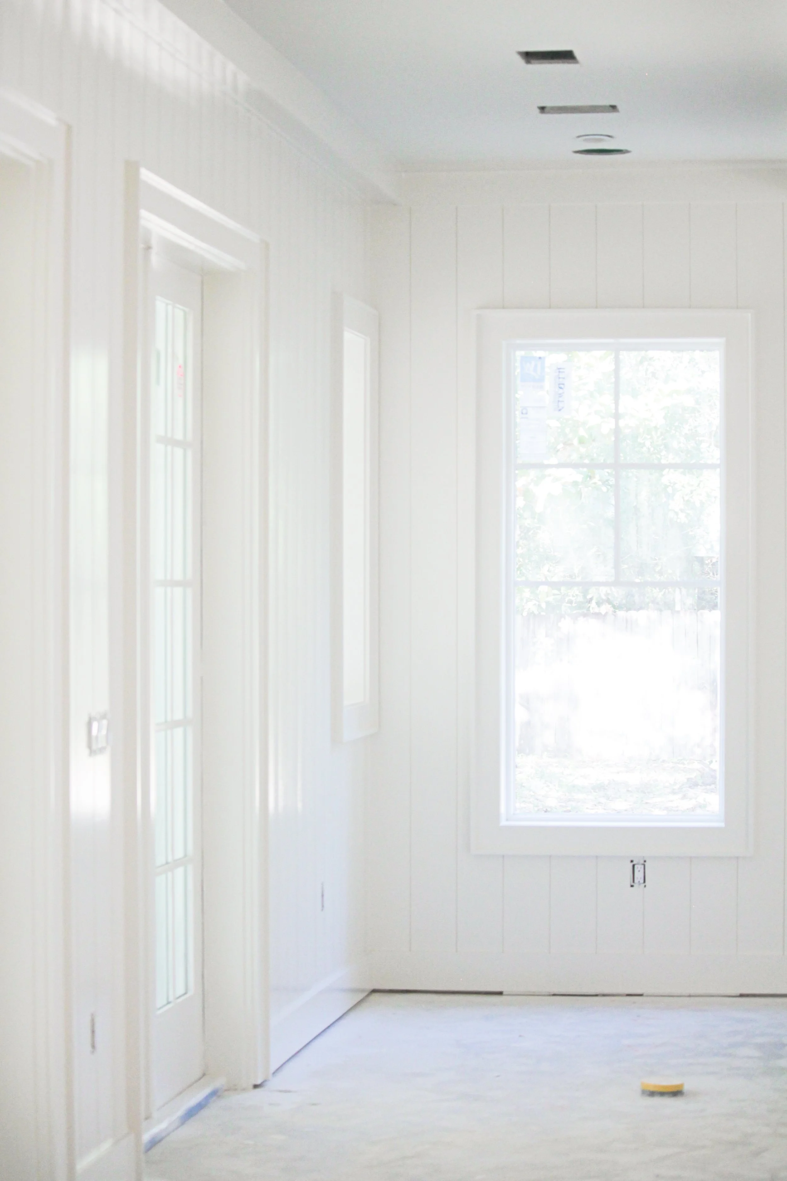 An interior room with white walls and ceiling. There is a large window on the right side and two smaller windows on the left. The room appears to be under construction or renovation, with electrical outlets and vents on the ceiling visible. The floor