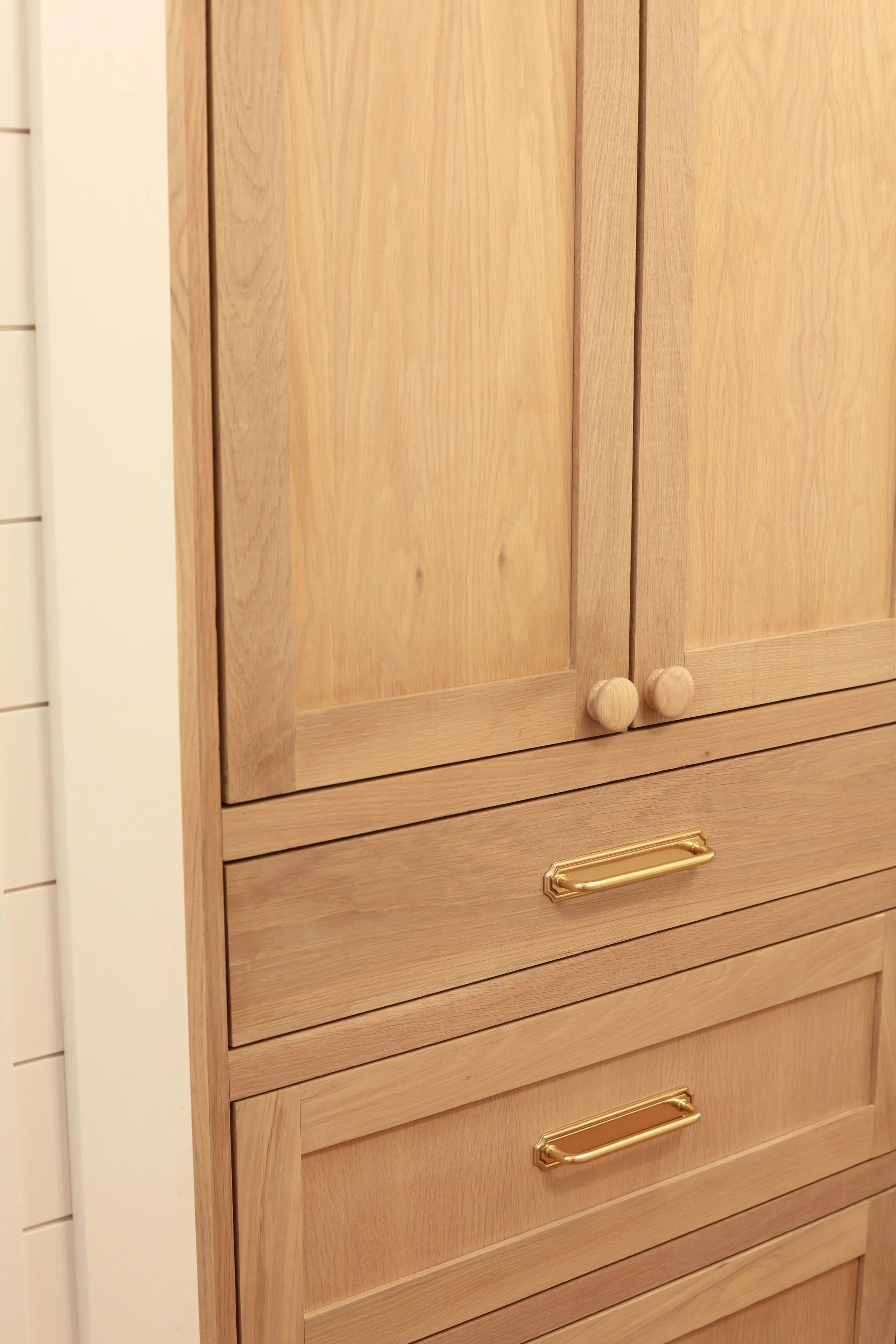 Close-up photo of a wooden kitchen cabinet with a gold-colored handle. The cabinet has a light wood finish with vertical slats.