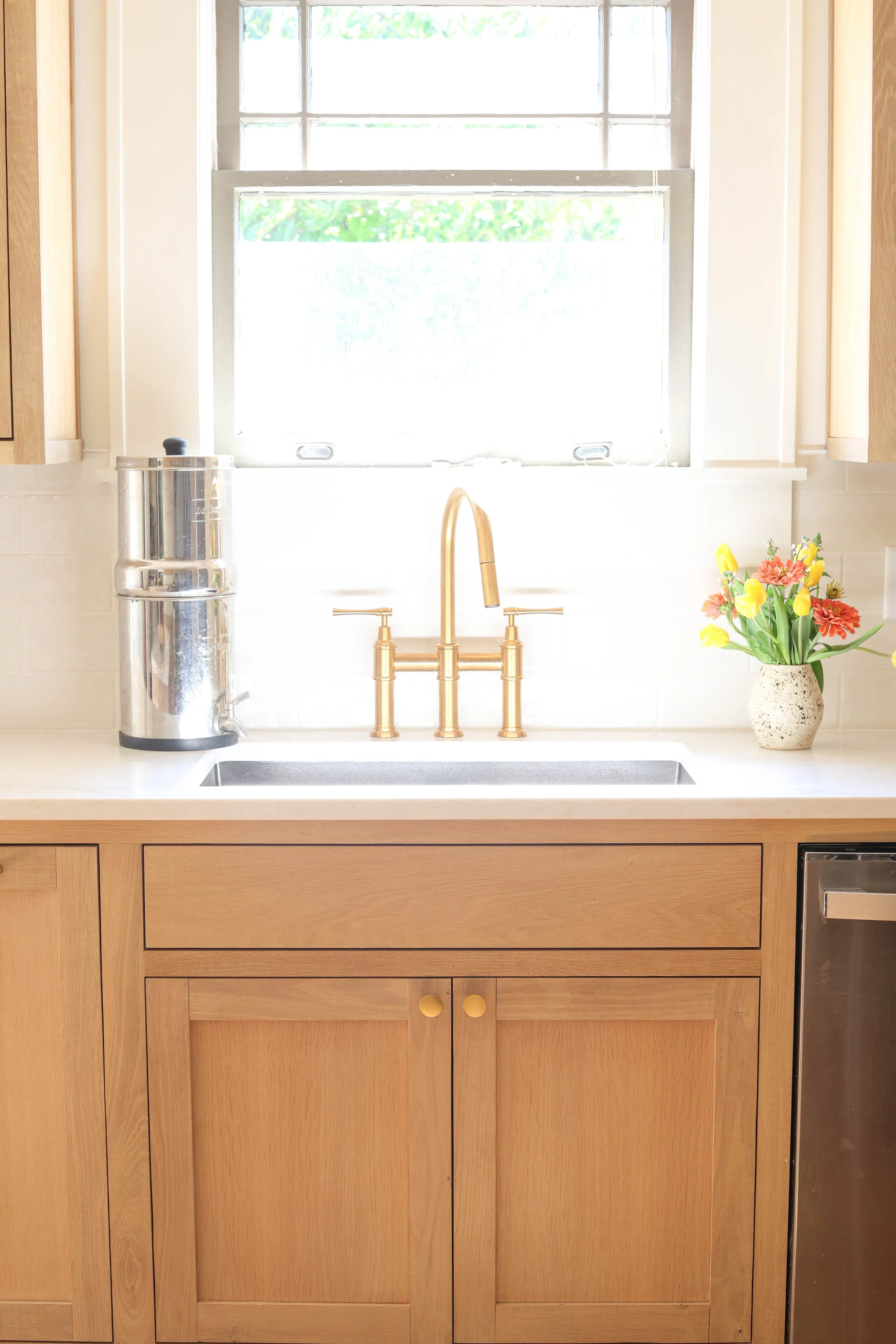 Kitchen sink with a gold faucet, a vase of flowers on the right, and a window above the sink with sunlight coming through, surrounded by light wood cabinets.