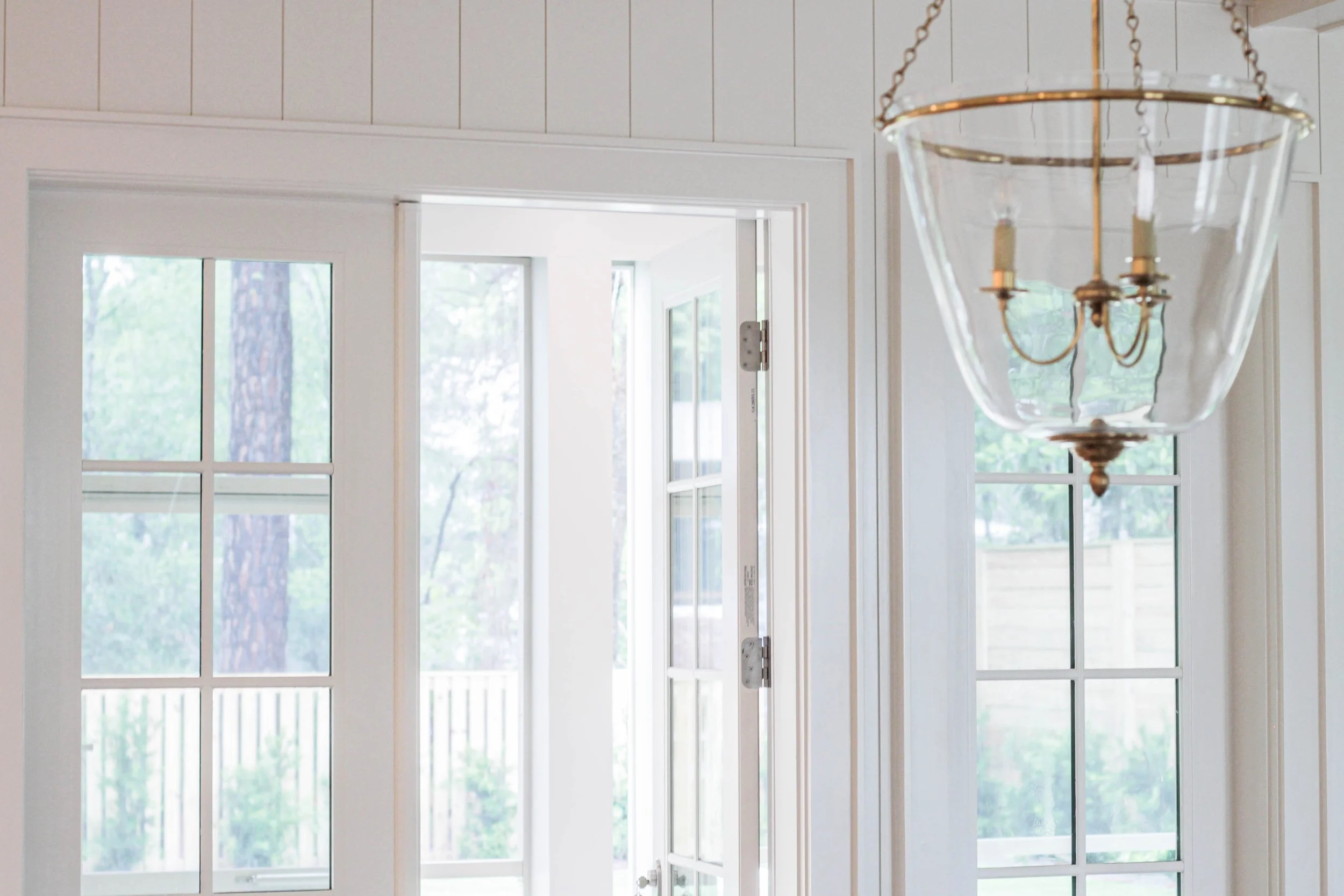 Close-up of a hanging glass light fixture with a brass chain, mounted near a large window with multiple panes showing a view of a tree outside.