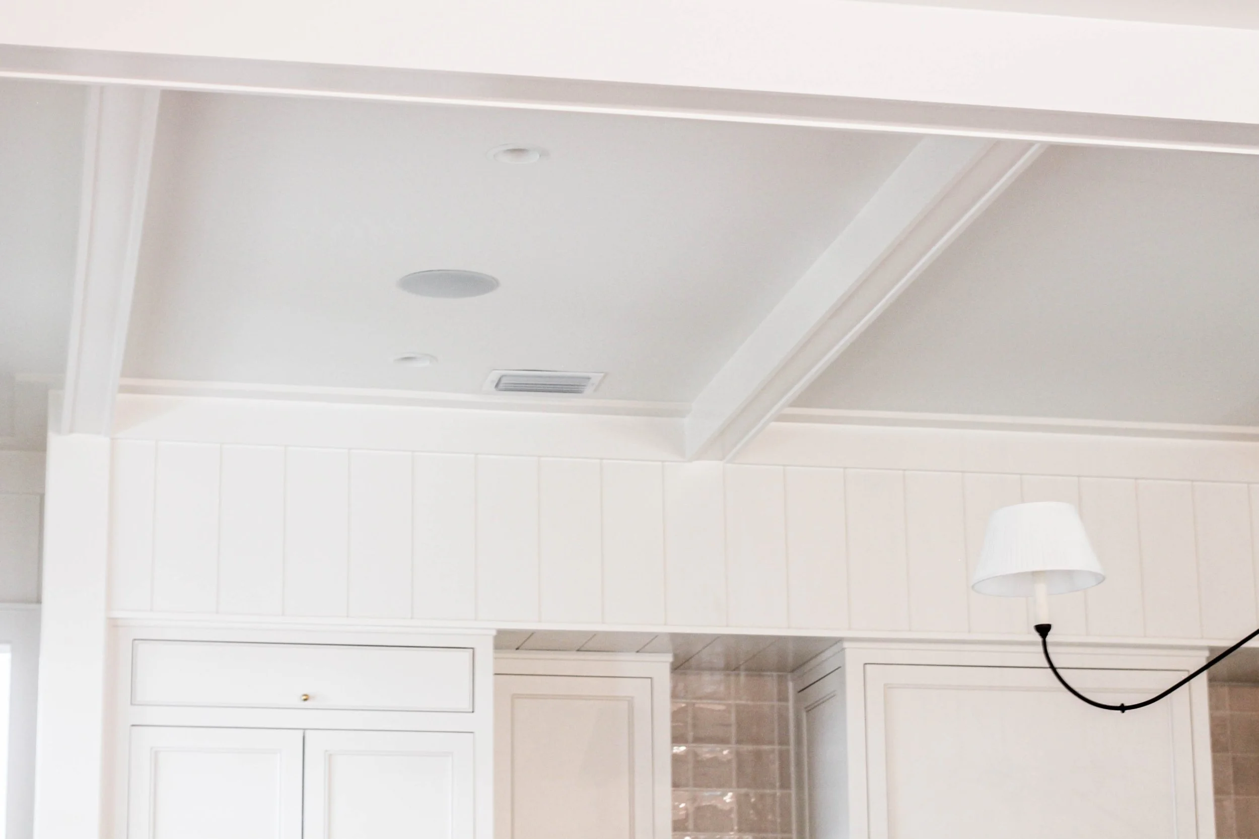 Part of a white ceiling with decorative molding, a recessed light, an air vent, a white wall with vertical paneling, a cabinet door, and a black lamp cord in a room with beige tiled flooring.