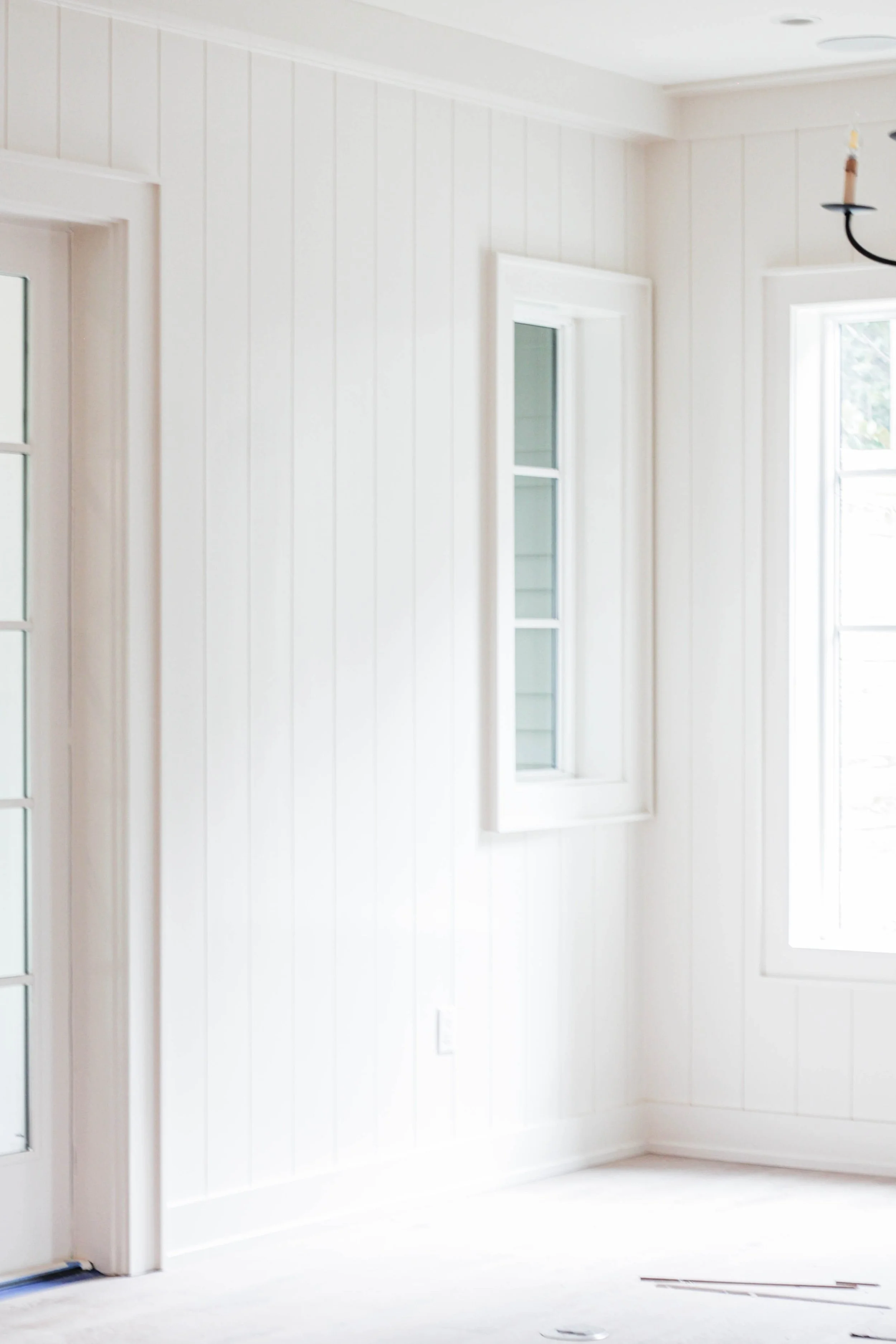 Empty room with white paneled walls, a window, and a black chandelier.