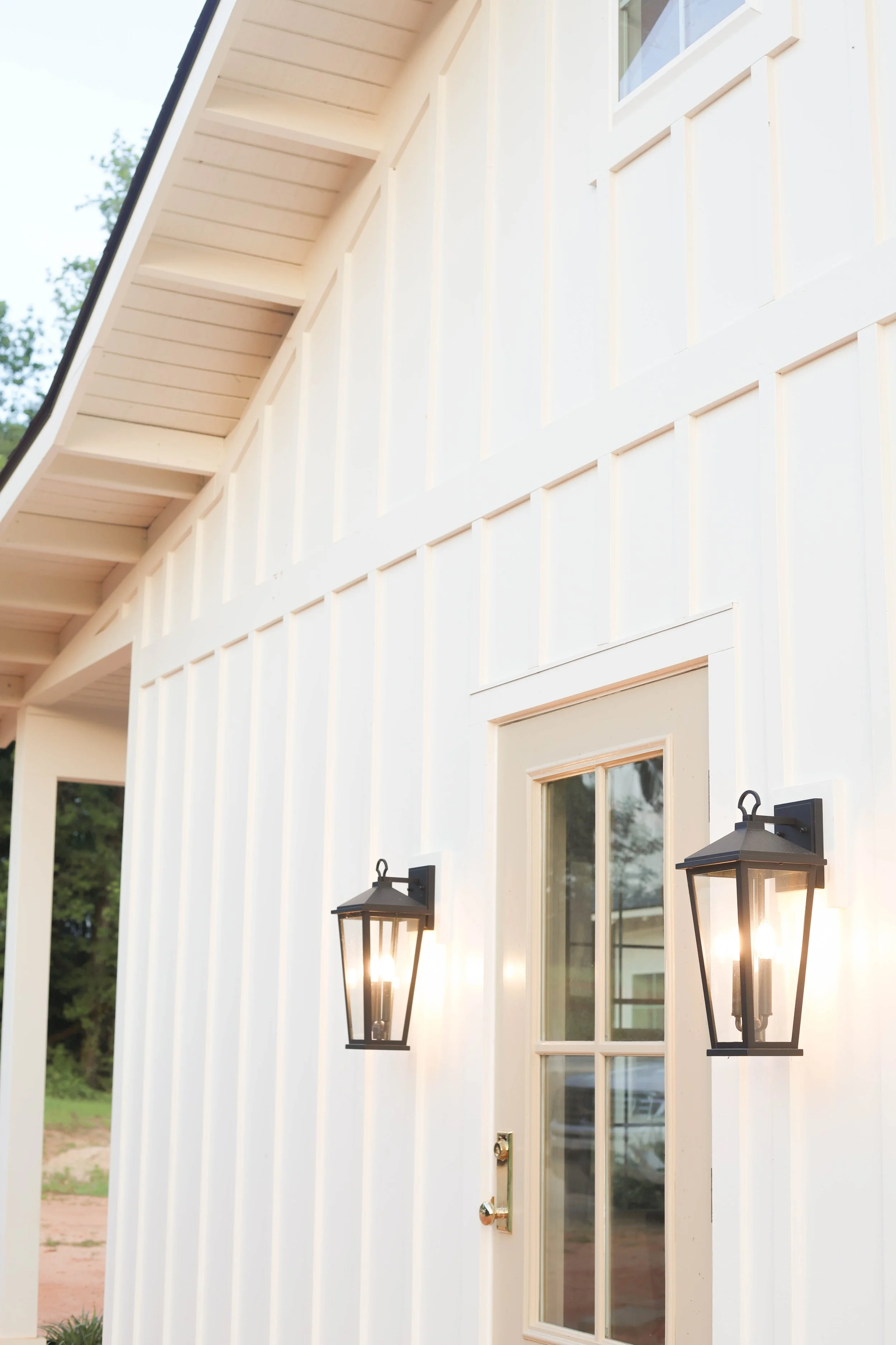 Close-up of a cream-colored exterior wall with a window, two black outdoor lanterns, and some greenery in the background.