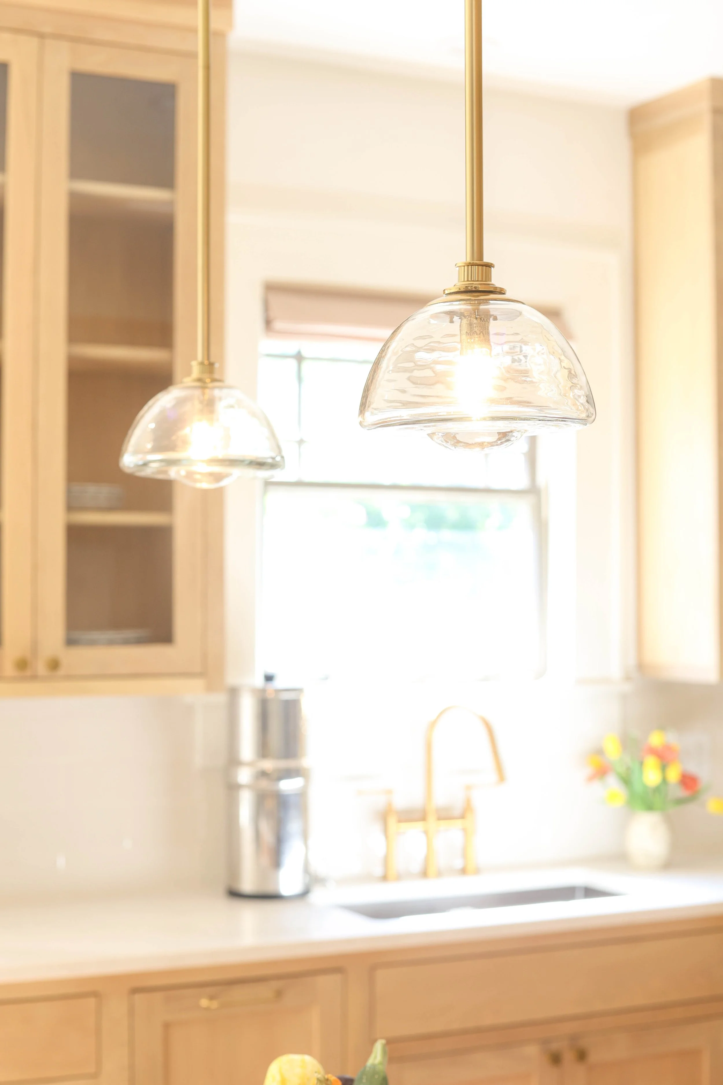Two glass pendant lights with brass fixtures hanging above a kitchen counter, with a background showing kitchen cabinets, a window, and a bouquet of flowers.