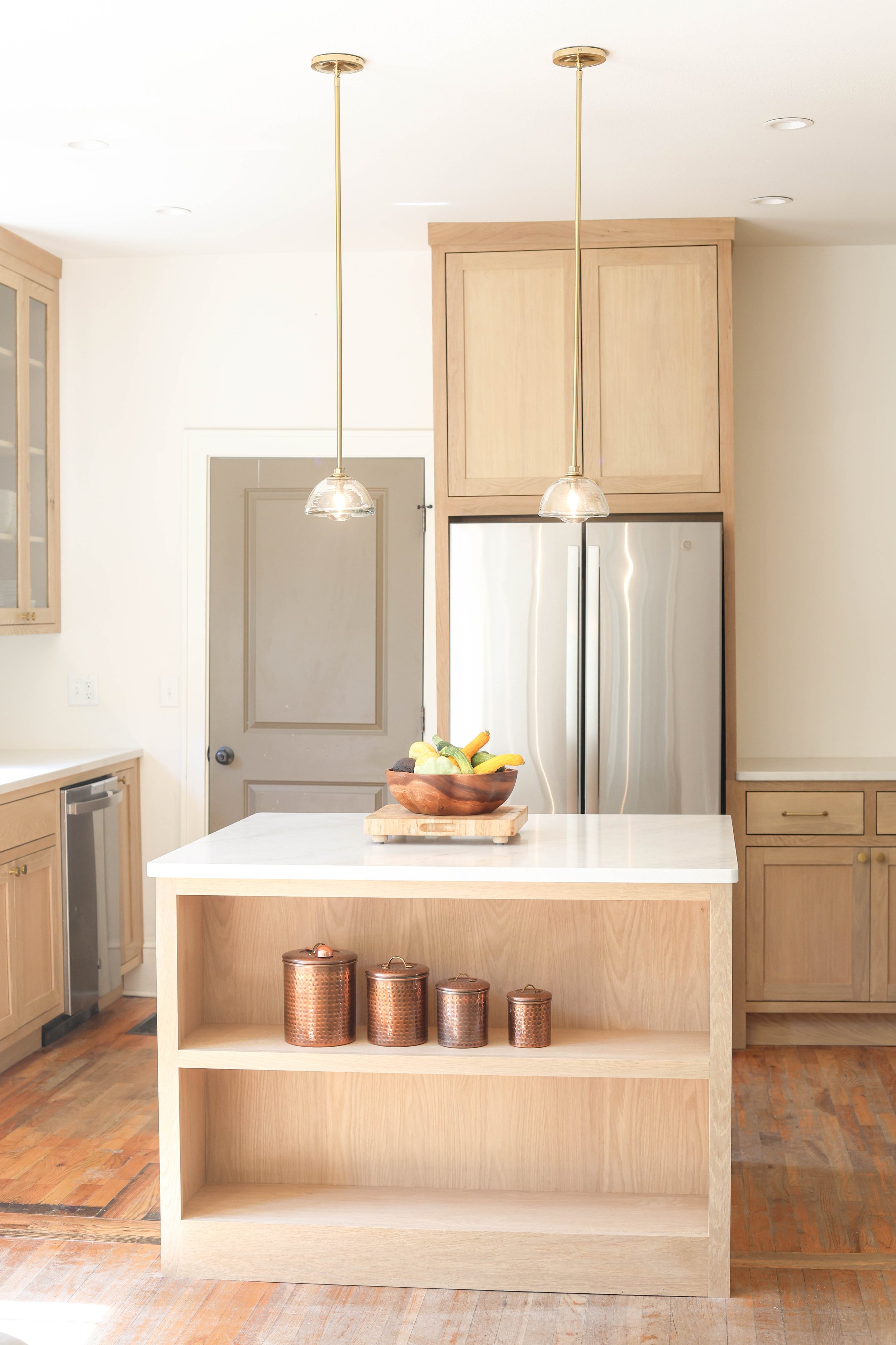 Modern kitchen with wood cabinetry, pendant lights, a white marble island with a wooden bowl of fruit, and copper canisters on open shelf.