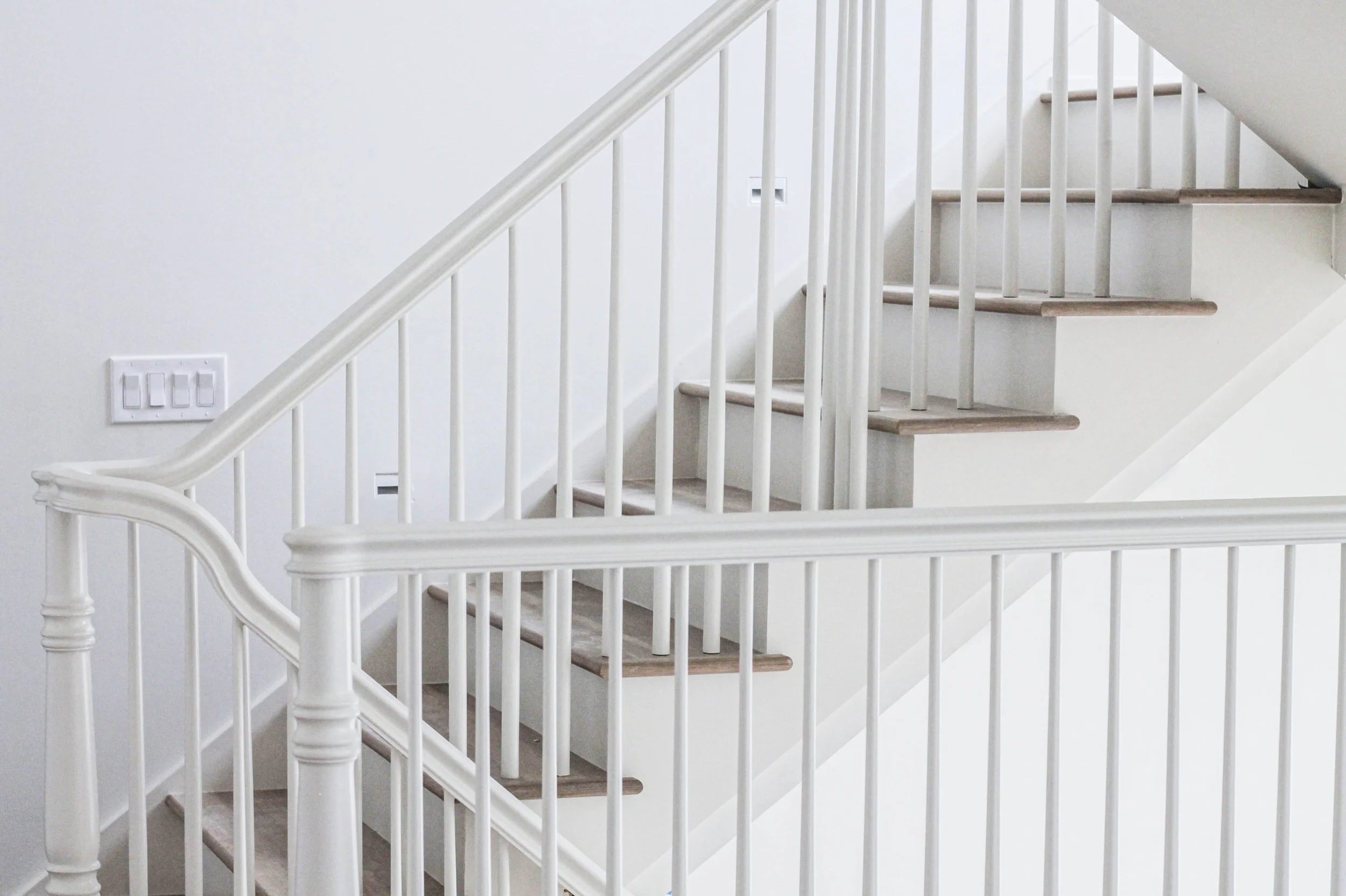 A white staircase with wooden steps and white railing, next to a white wall with a light switch and an electrical outlet.
