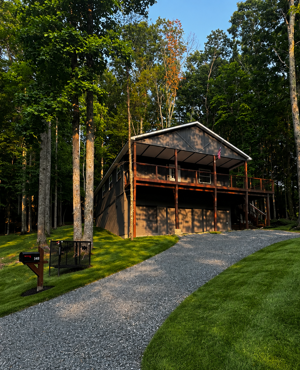 A modern house on a wooded hill with a gravel driveway, surrounded by green grass and trees, under a blue sky.