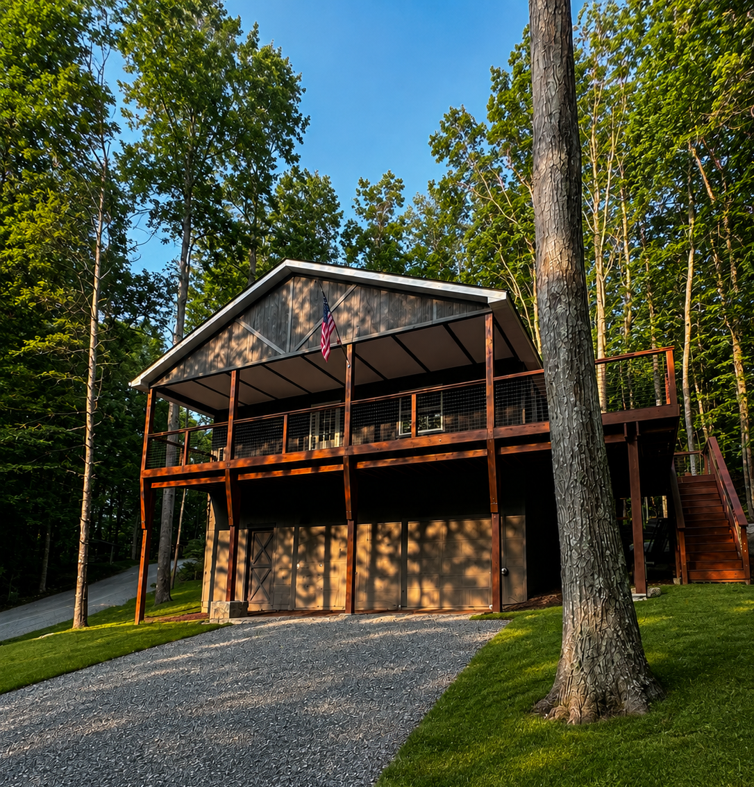 A two-story house with a garage on the ground floor, wooden exterior, and a balcony on the upper level, surrounded by tall trees and green grass, with an American flag on the balcony, during sunset.