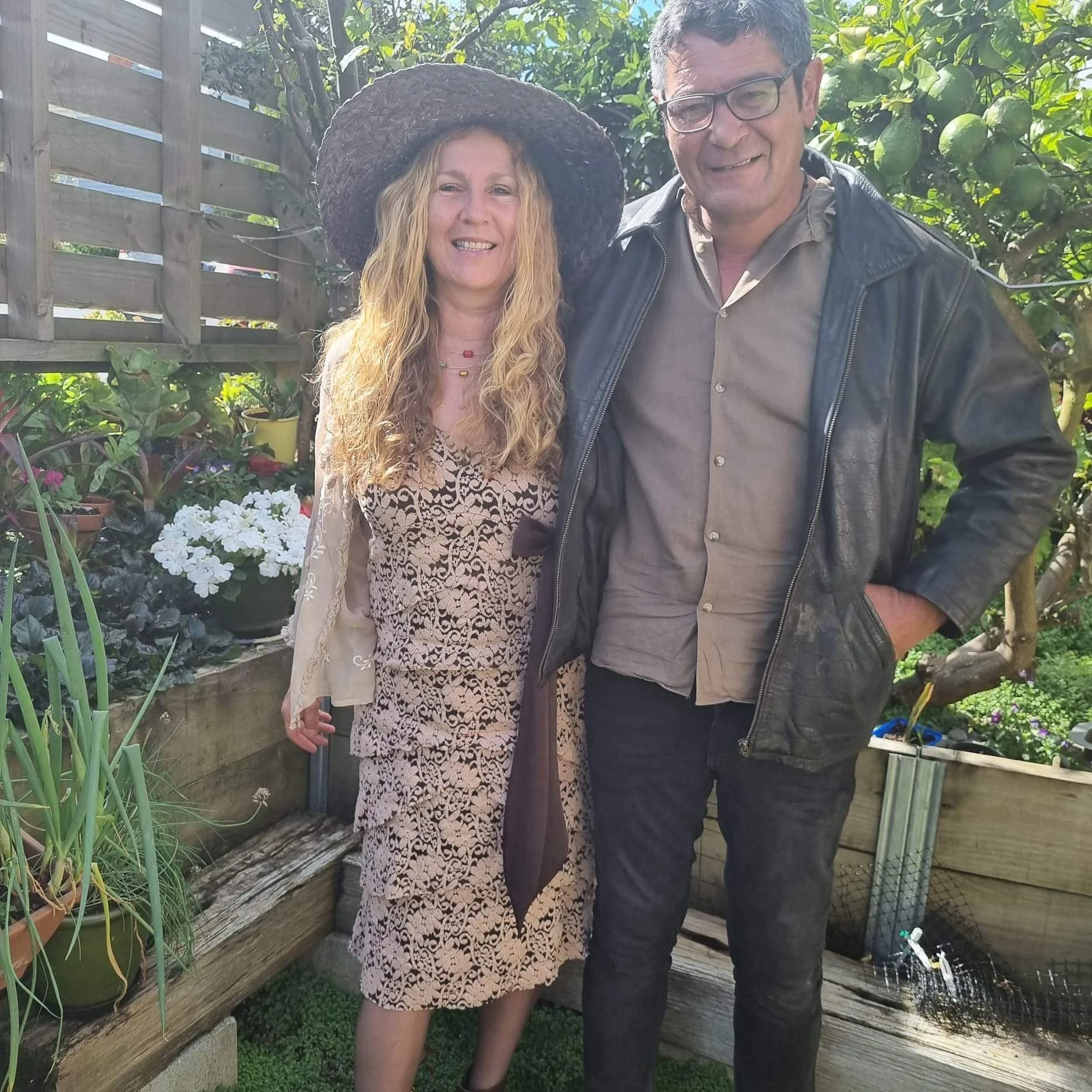A woman with long curly blonde hair wearing a wide-brimmed hat and patterned dress, standing next to a man with short hair, glasses, and a dark jacket, in a garden with potted plants and flowering bushes.