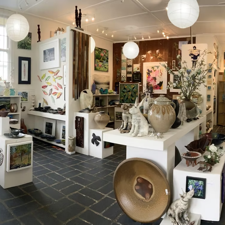Interior of an art gallery or store with various artworks, ceramics, sculptures, and decorative items on display, lit by hanging paper lantern lights.