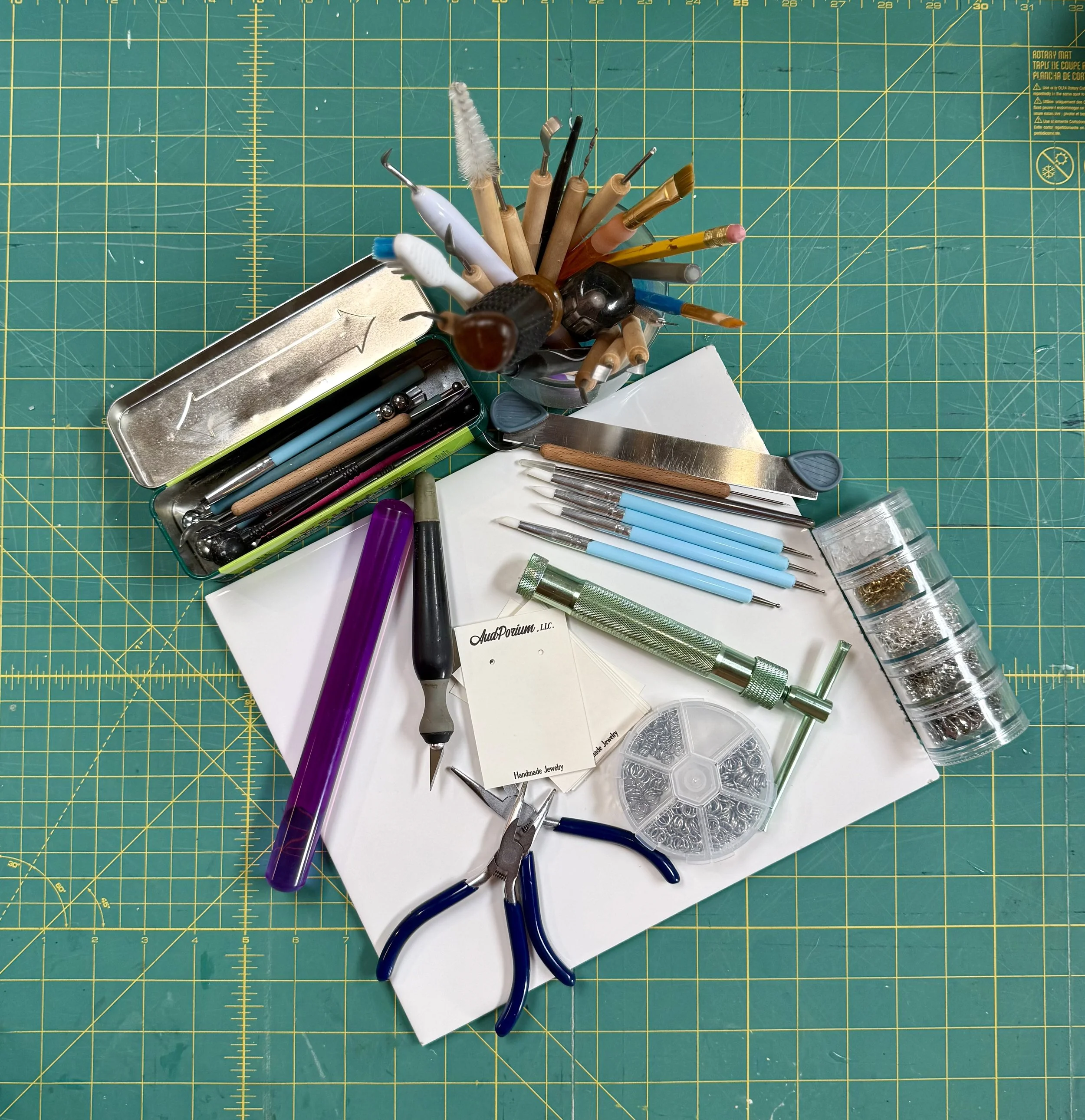 A green cutting mat with yellow grid lines. On the mat, there is a cup holding various tools and brushes, a metal tray with a white arrow drawn on it, and several pens, markers, and tools scattered around, along with small containers of beads and metallic findings, likely for jewelry making.