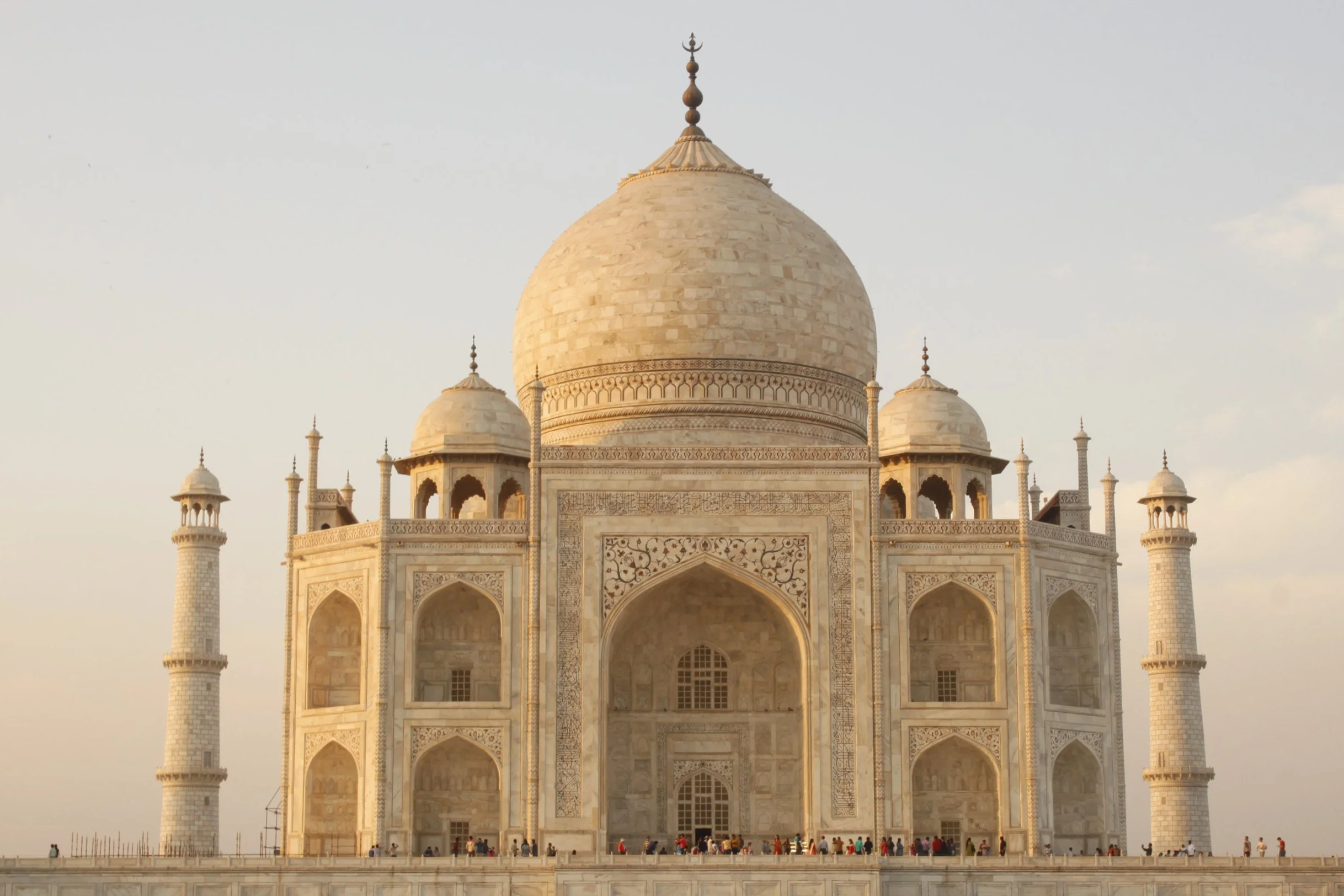 The Taj Mahal, a white marble mausoleum with a large central dome, four smaller domes, and four minarets, with visitors on the platform in front during daytime.
