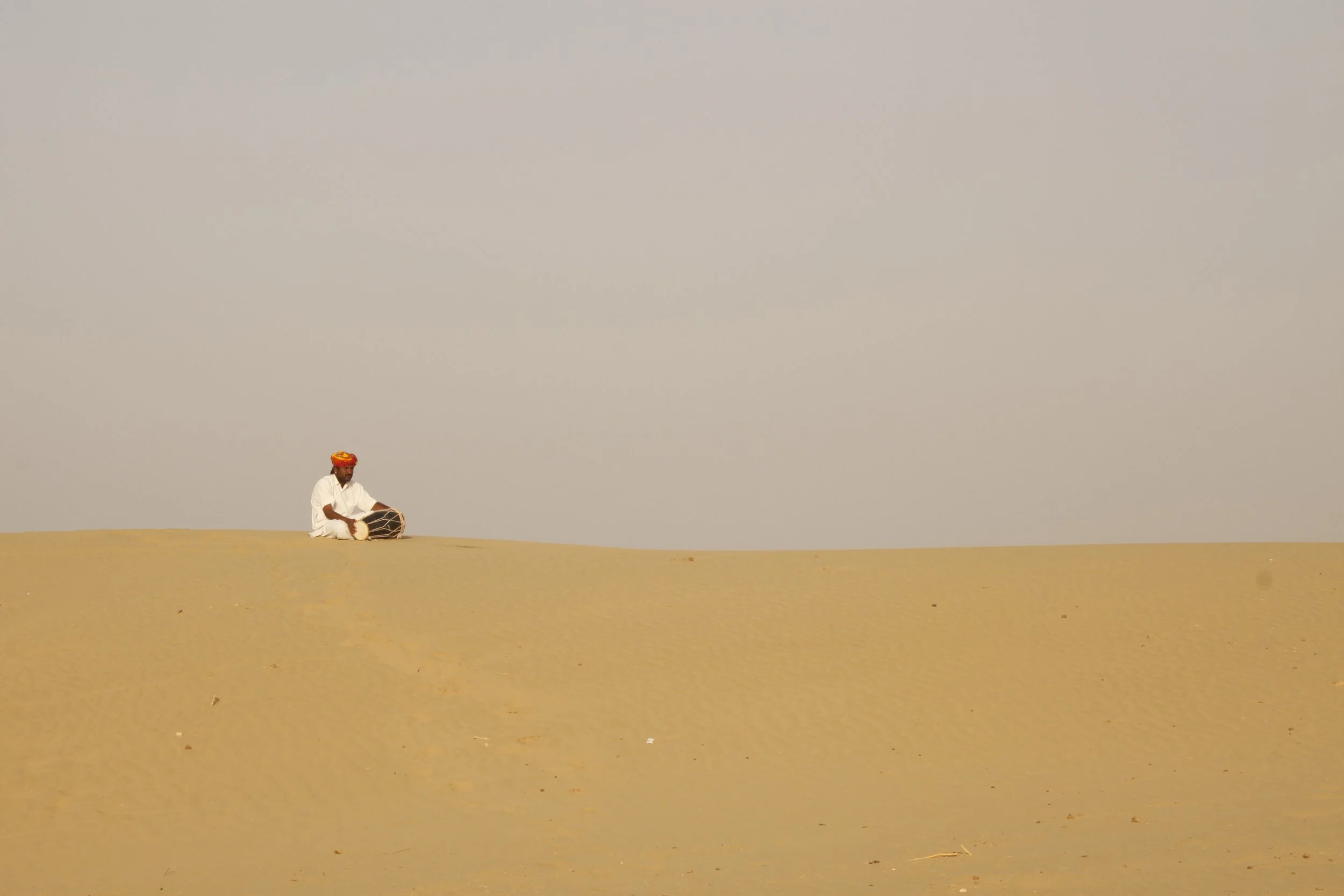 A man dressed in white sitting on a sandy desert playing a drum with a colorful head covering.