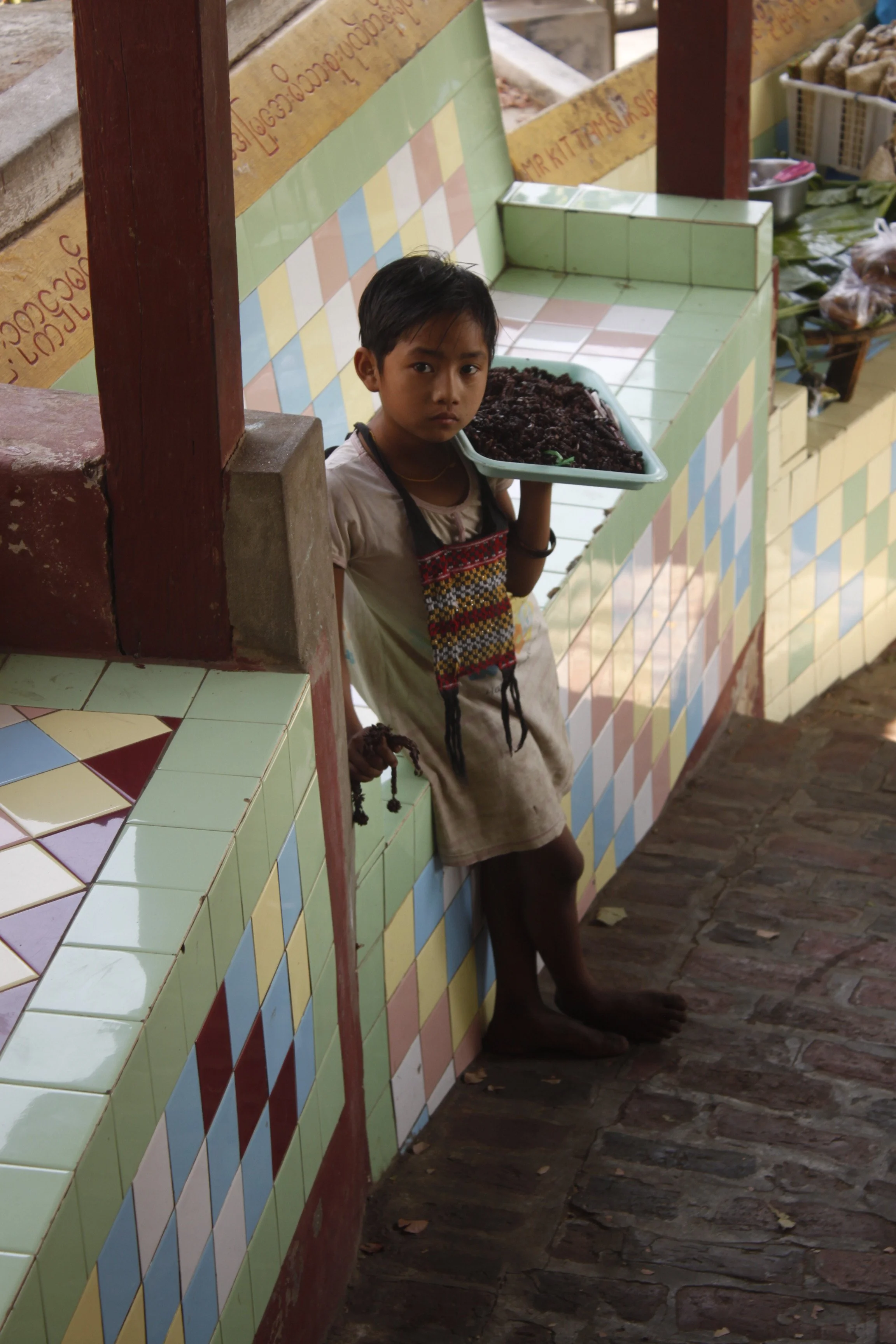 Young boy with dark hair, standing barefoot against a colorful tiled counter, holding a tray of dark food, in a market or shop setting.