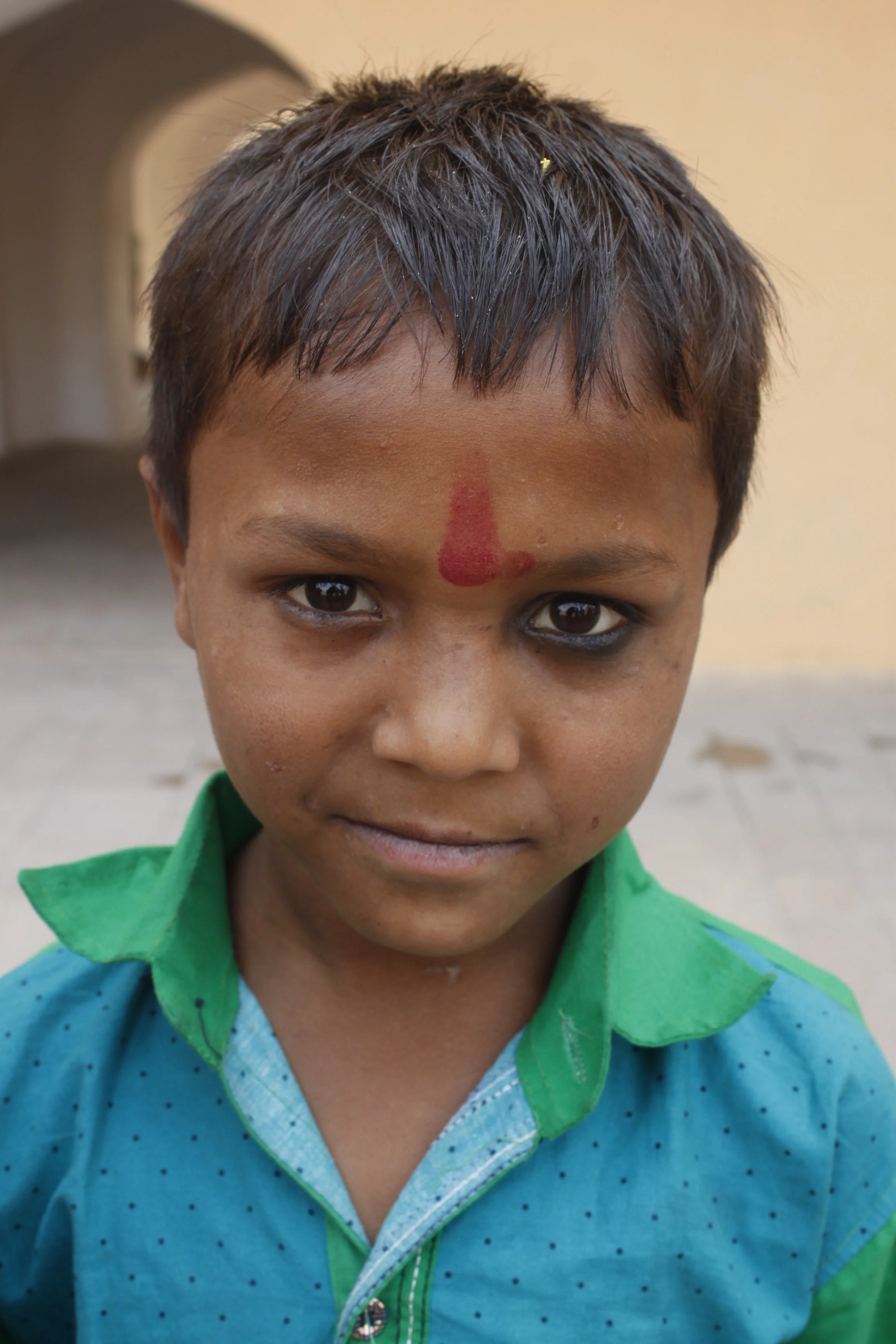 A young boy with short dark hair and dark eyes, wearing a colorful jacket, has red tilak on his forehead, and is looking directly at the camera with a slight smile.