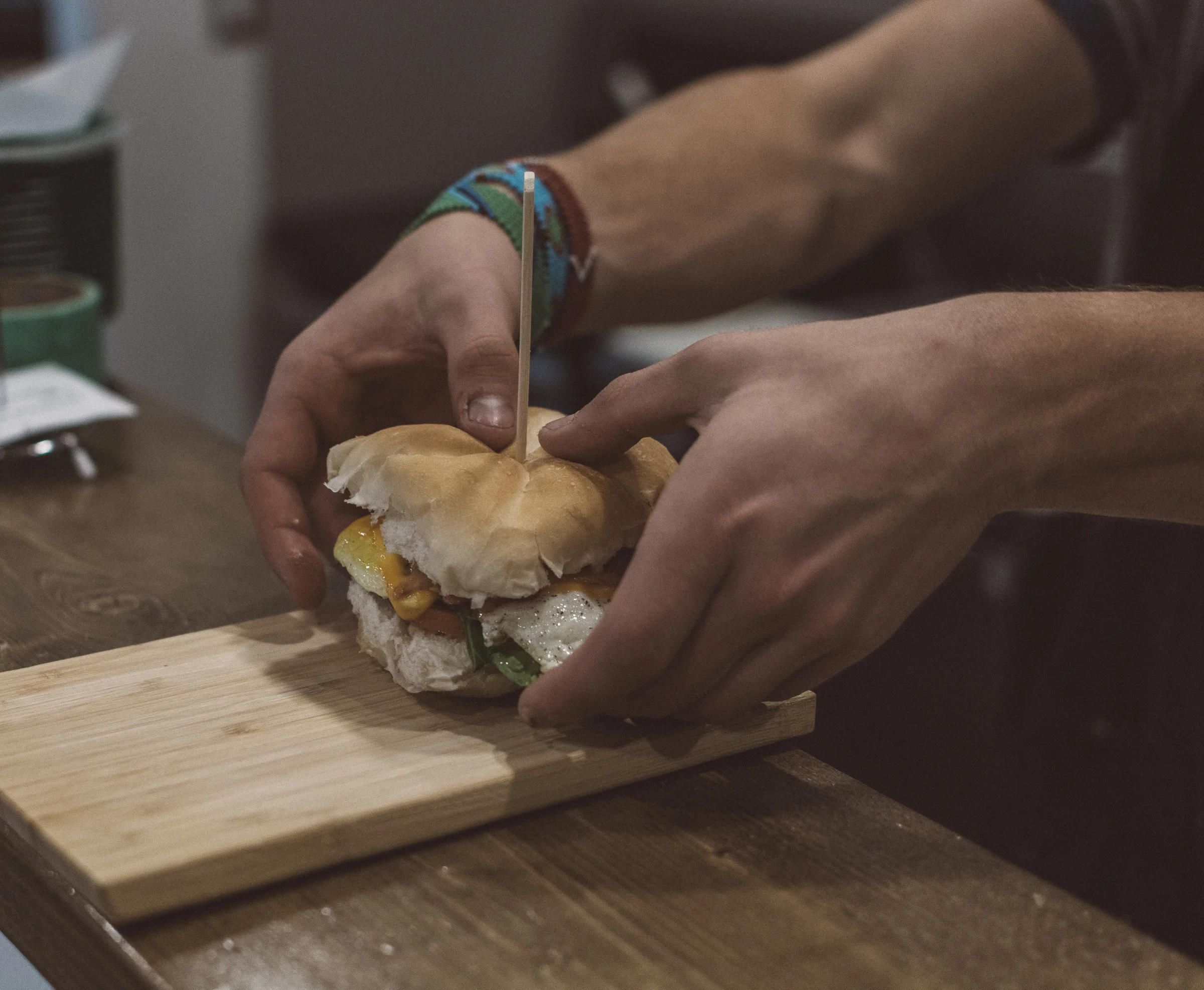 A person assembling a sandwich or burger on a wooden cutting board, with a toothpick inserted into the sandwich. The person's hands are visible, and they are wearing a colorful, patterned wristband.