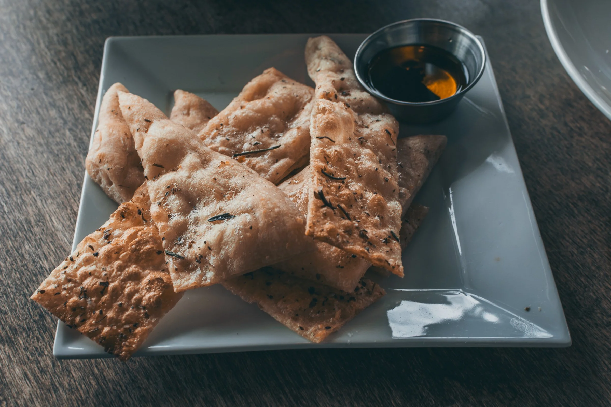 Three pieces of crispy flatbread sprinkled with herbs on a white square plate, accompanied by a small black dish of dipping sauce, set on a dark wooden table.