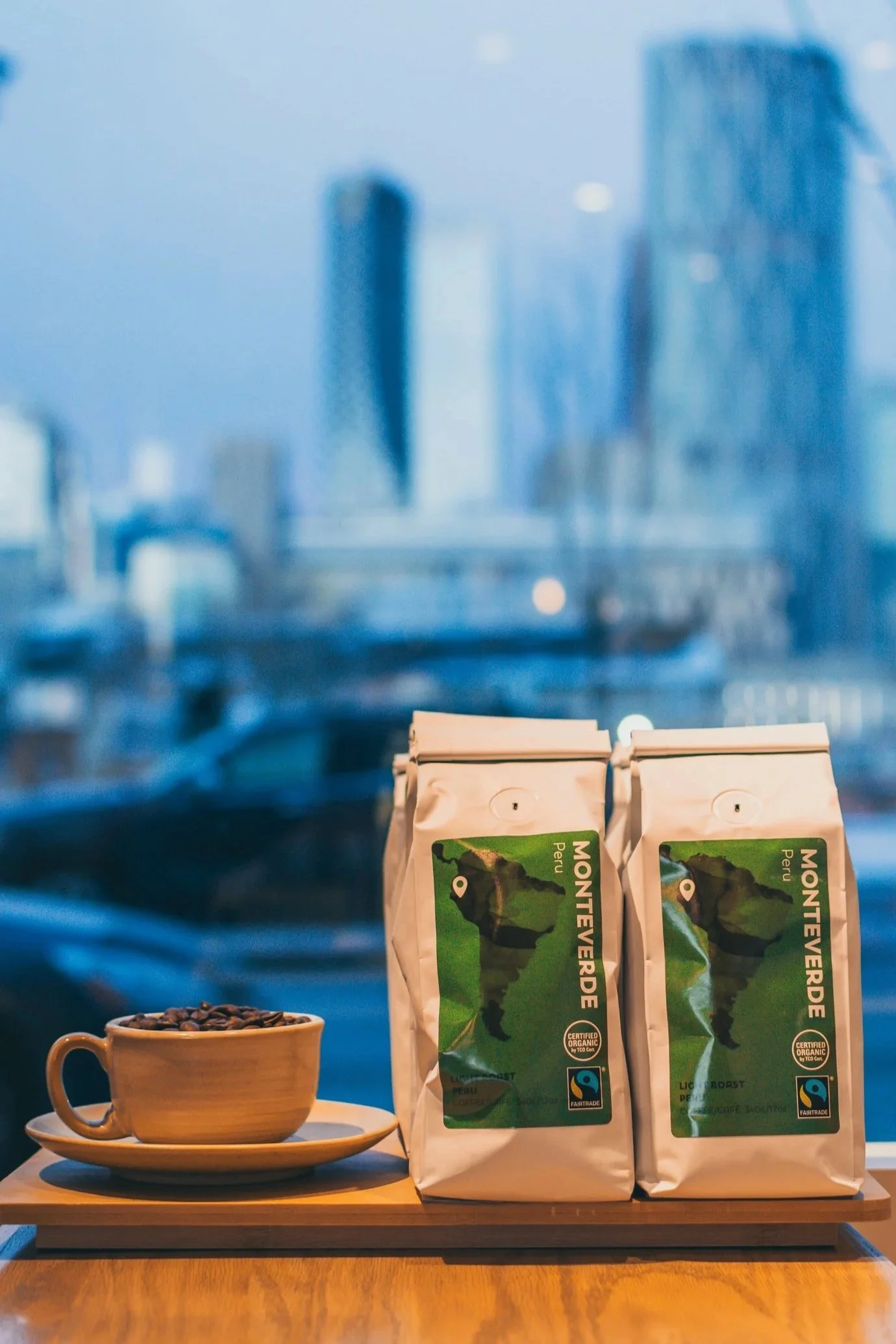 Three bags of Fairtrade certified organic coffee with a scenic picture of a mountain on the label, placed on a wooden table, with a ceramic cup of coffee and a saucer filled with coffee beans in front of them.