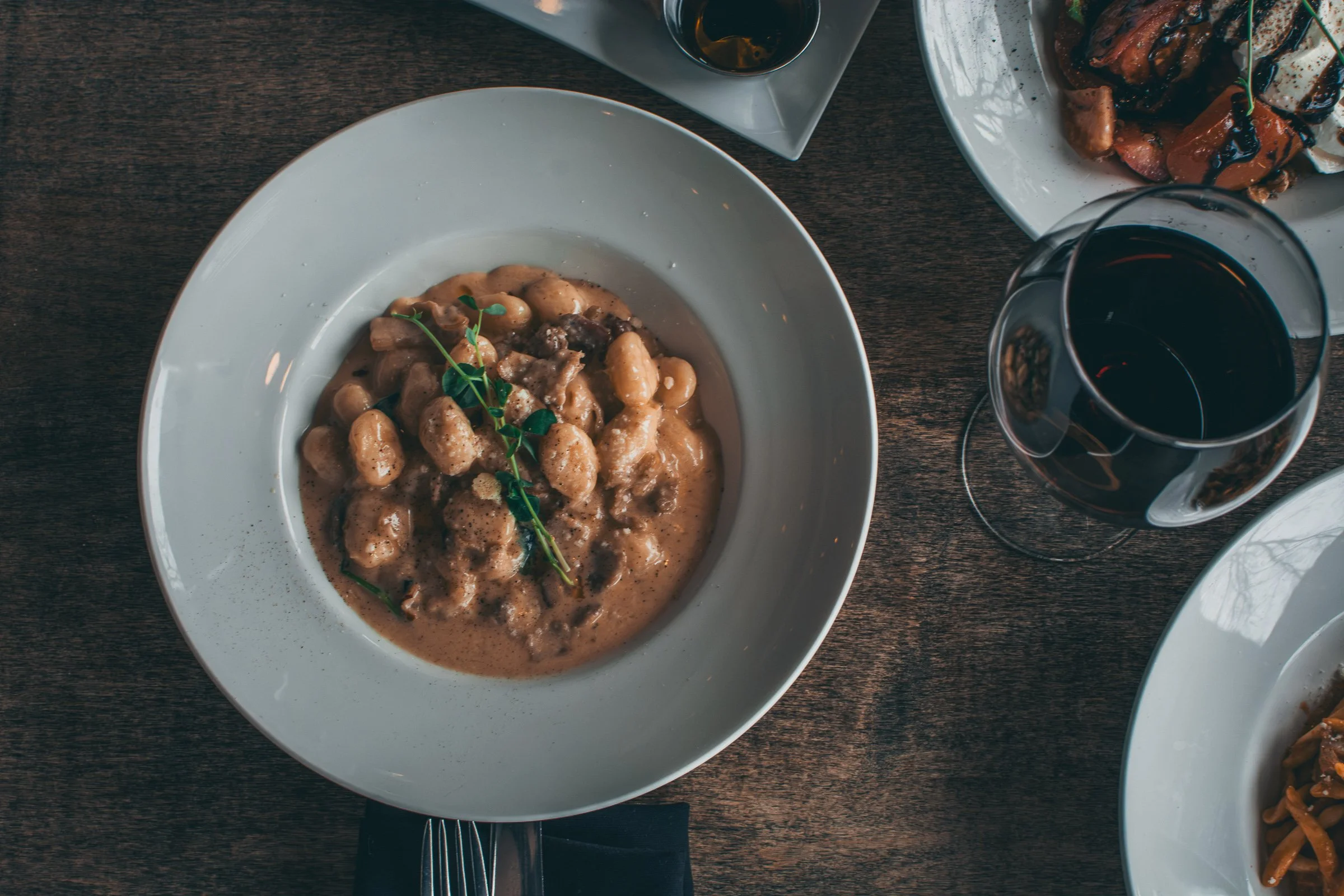 A white plate of gnocchi in a cream sauce garnished with a sprig of fresh herbs on a dark wooden table, with a glass of red wine and other dishes partially visible around it.