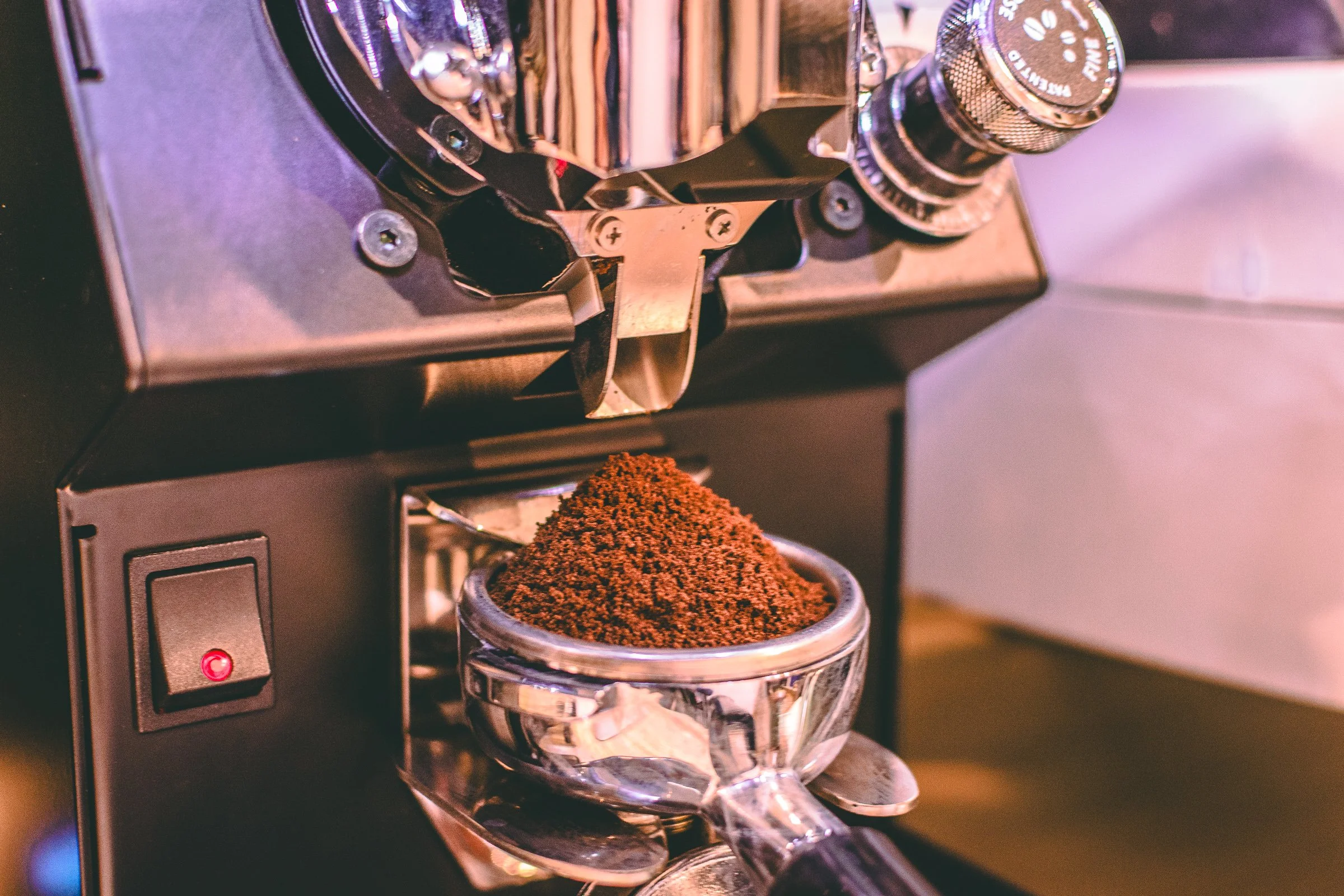 Close-up of a coffee grinder with ground coffee in a transparent glass container underneath, and part of the grinder's metallic lid visible.