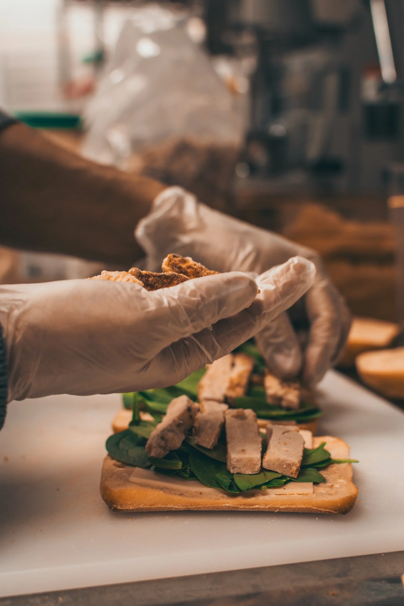 Close-up of a person wearing gloves preparing a sandwich or wrap with slices of cooked meat, lettuce, and bread, on a wooden cutting board in a kitchen.