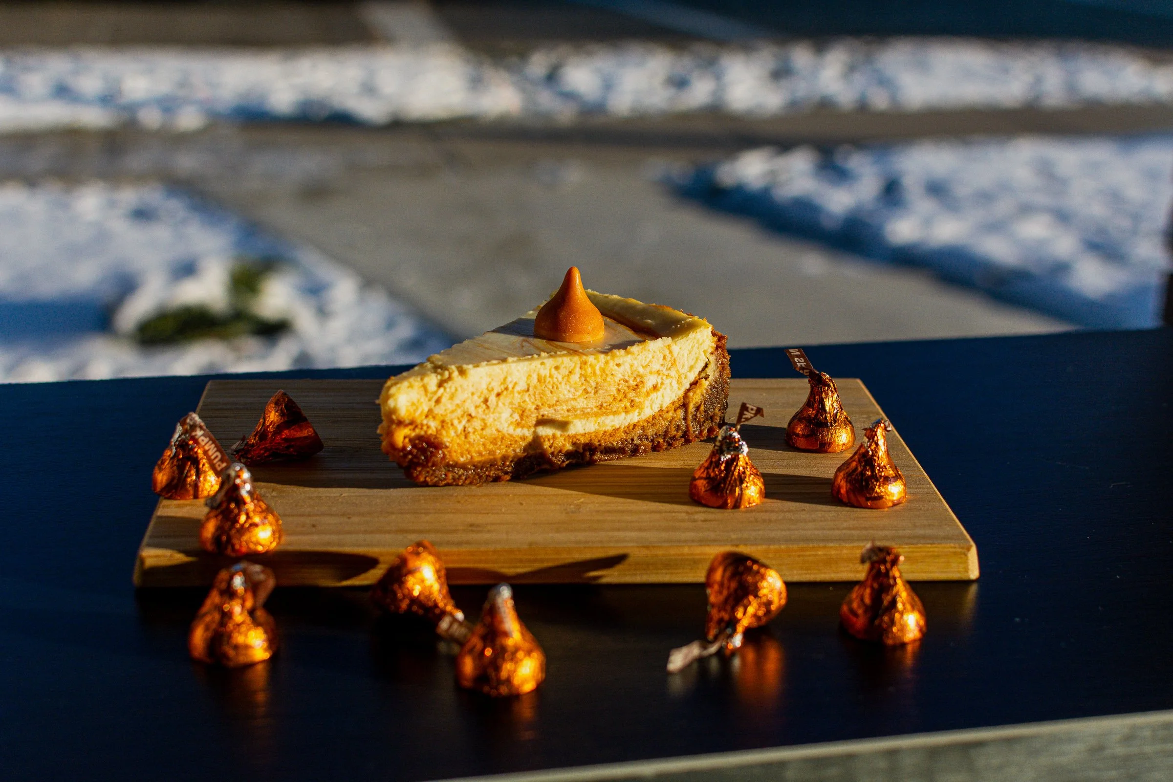 A slice of cheesecake with a caramel drizzle on top, decorated with small, shiny, gold and copper-colored wrappers surrounding it, on a wooden serving board, with a snowy outdoor scene in the background.