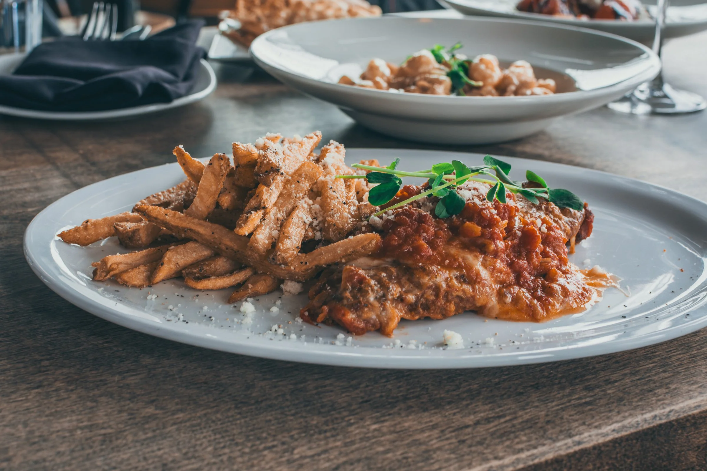 Plate of fried French fries topped with grated cheese and garnished with green microgreens, served with a side of lasagna with tomato sauce, served on a white plate.
