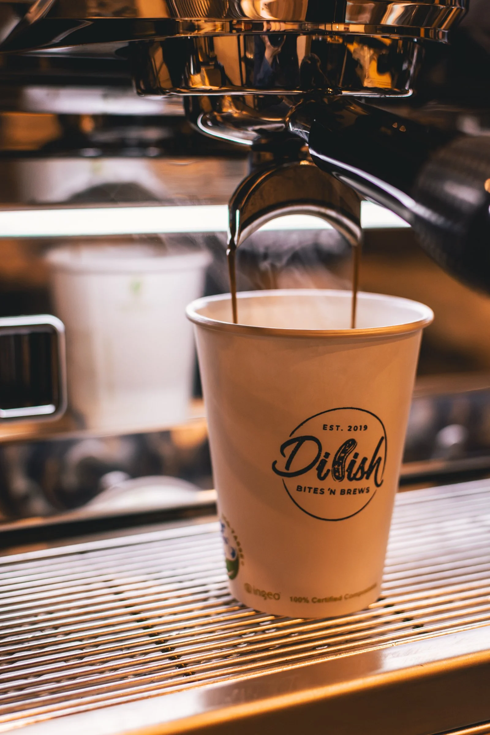 Coffee being brewed from espresso machine into a paper cup with a logo that says 'D'lish' and the phrase 'Bites n Brews'.