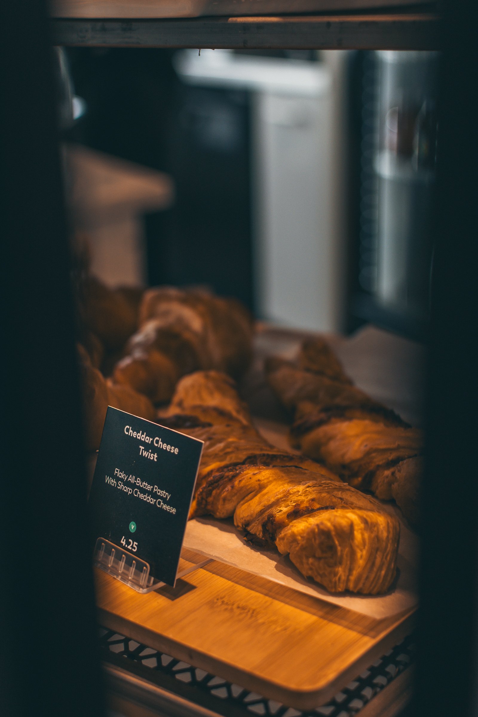 A tray of croissants, with a sign that reads 'Cheddar Cheese Twist' and describes it as flaky all-butter pastry with sharp cheddar cheese, priced at $4.25, in a bakery display case.