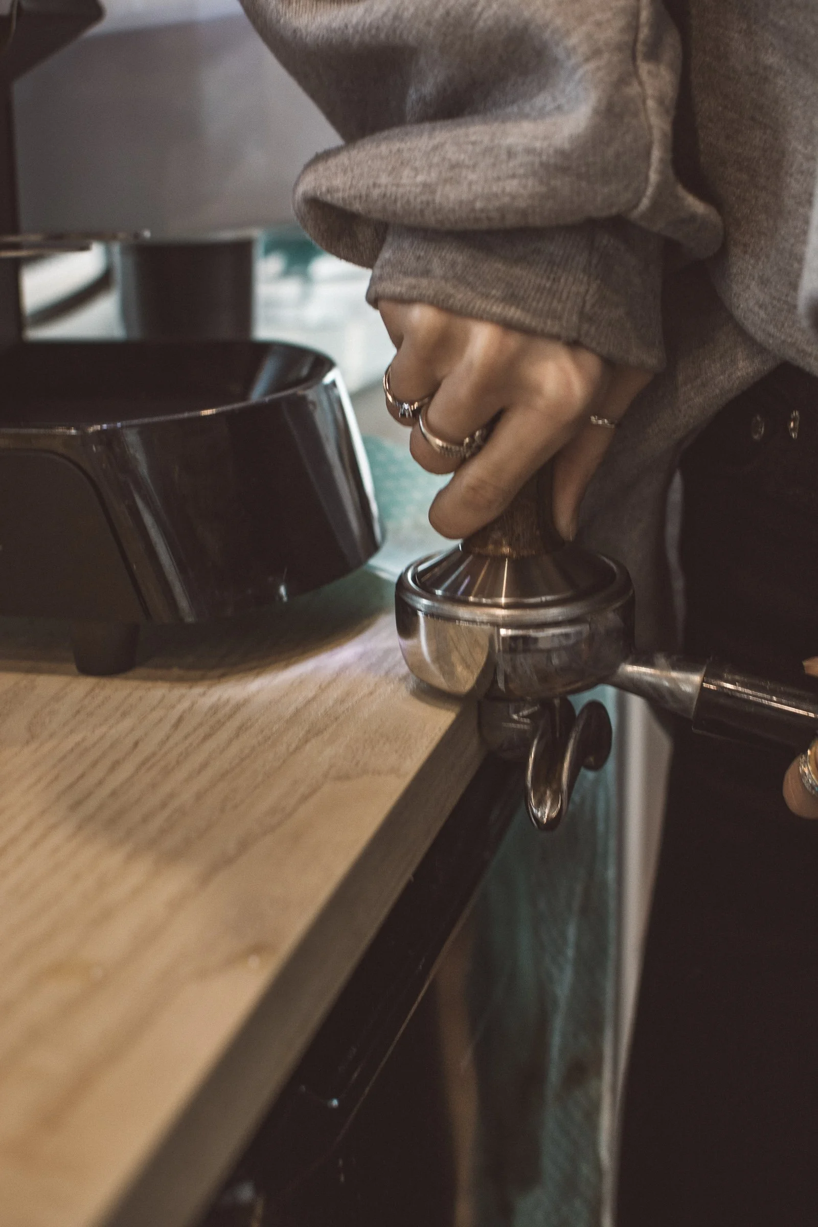 A person wearing a brown long-sleeve shirt using a metallic manual coffee grinder on a wooden surface.