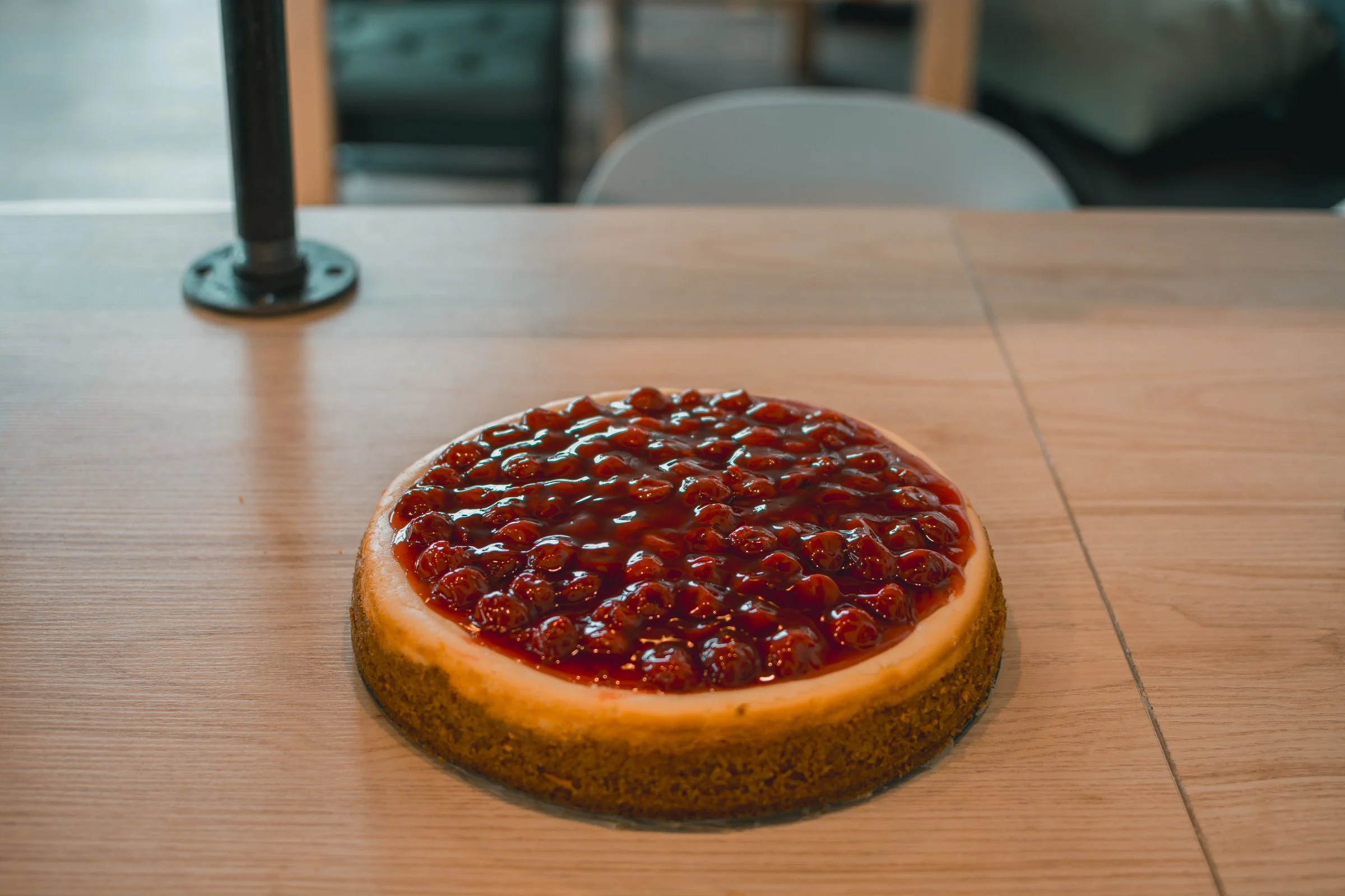 A slice of cherry cheesecake with a crumbly crust and cherry topping on a wooden table in a cafe or restaurant setting.