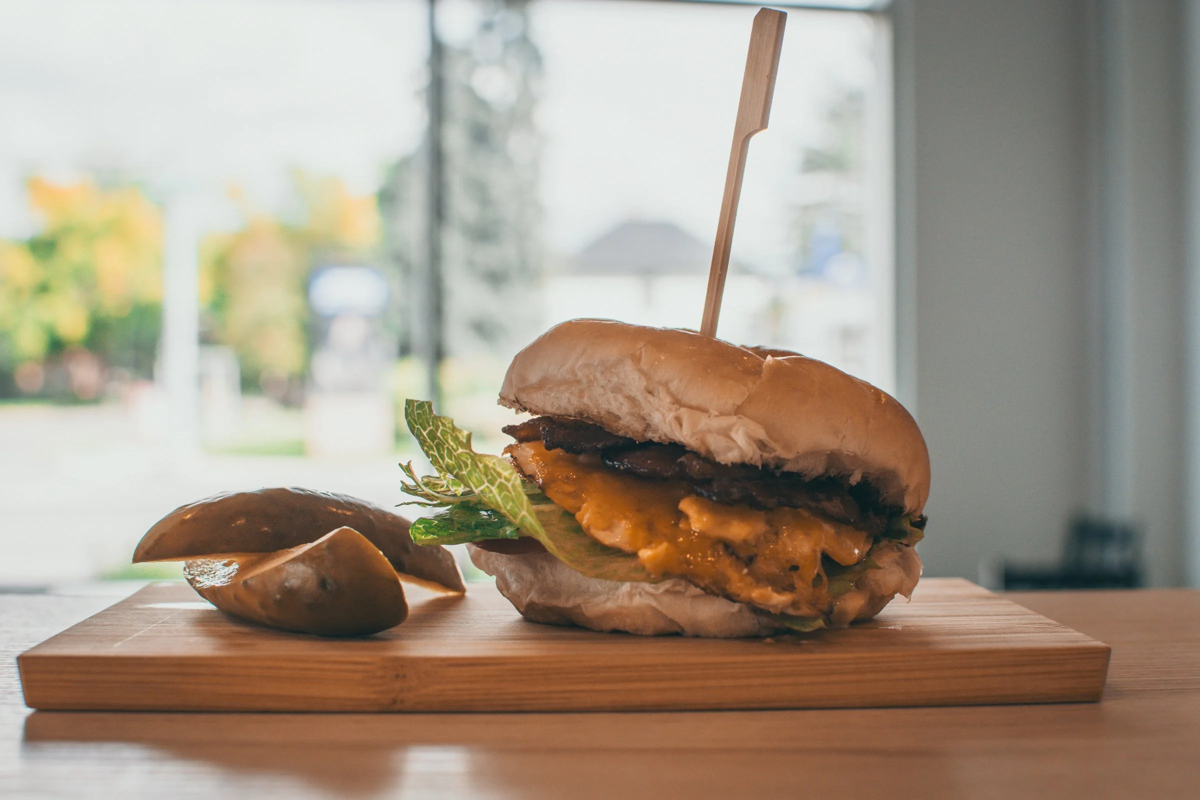 Close-up of a burger with cheese, lettuce, and a toothpick, placed on a wooden cutting board with a blurred window in the background.