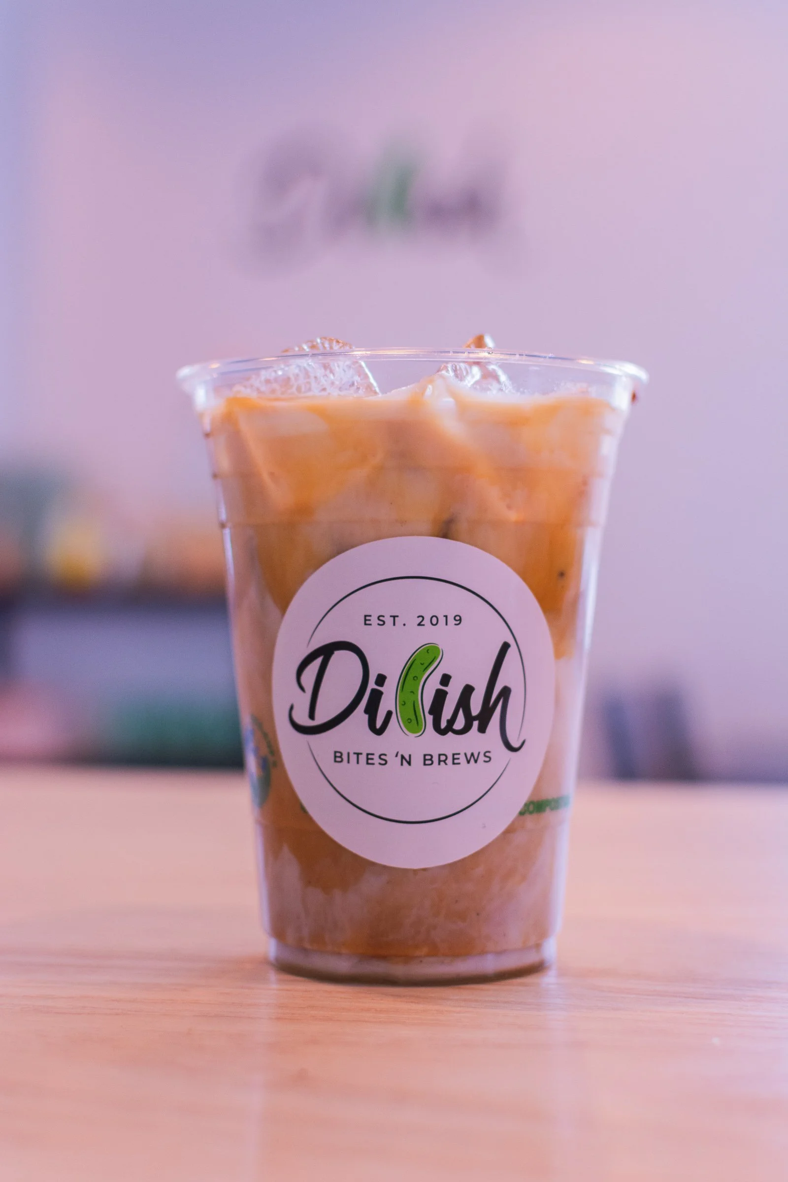 A plastic cup of iced coffee with a label that says 'Dilish' and 'Bites 'n Brews,' placed on a wooden surface with a blurred background.