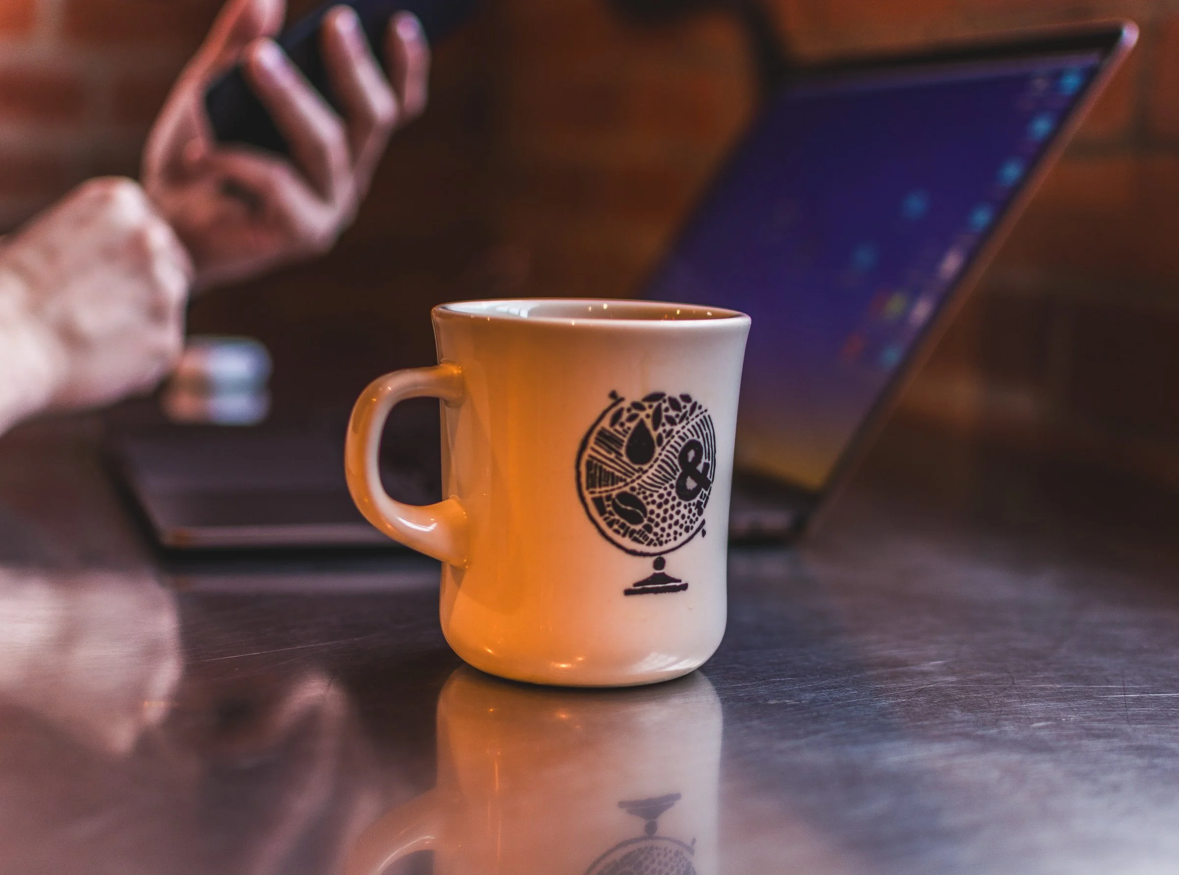 A white ceramic mug with a black graphic of a stylized face and earthenware design, sitting on a reflective dark wooden surface. In the background, a person's hand is reaching toward a tablet with a blurred screen.