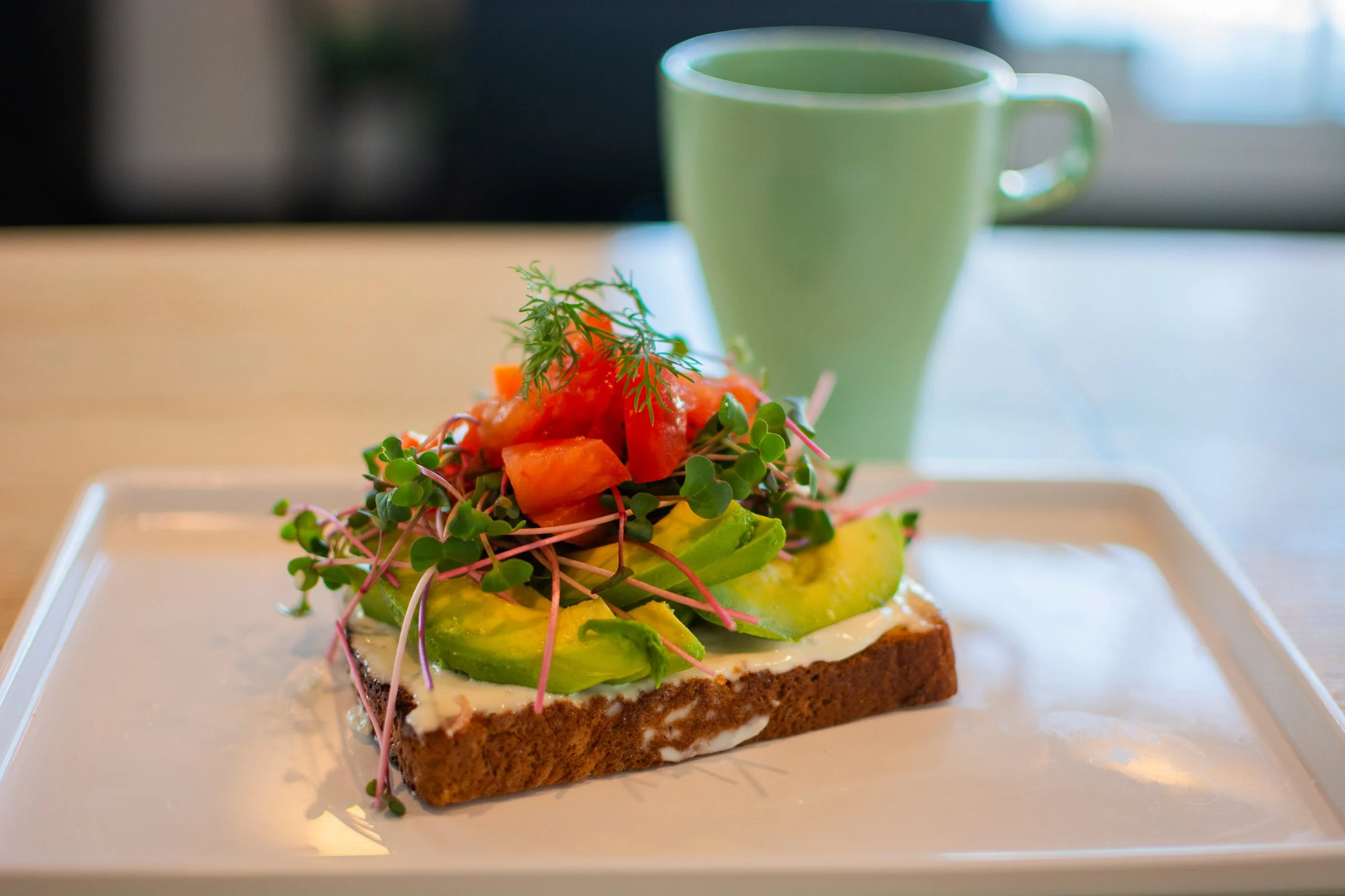 A slice of avocado toast topped with microgreens, diced tomatoes, and herbs on a white plate with a light-colored mug in the background.