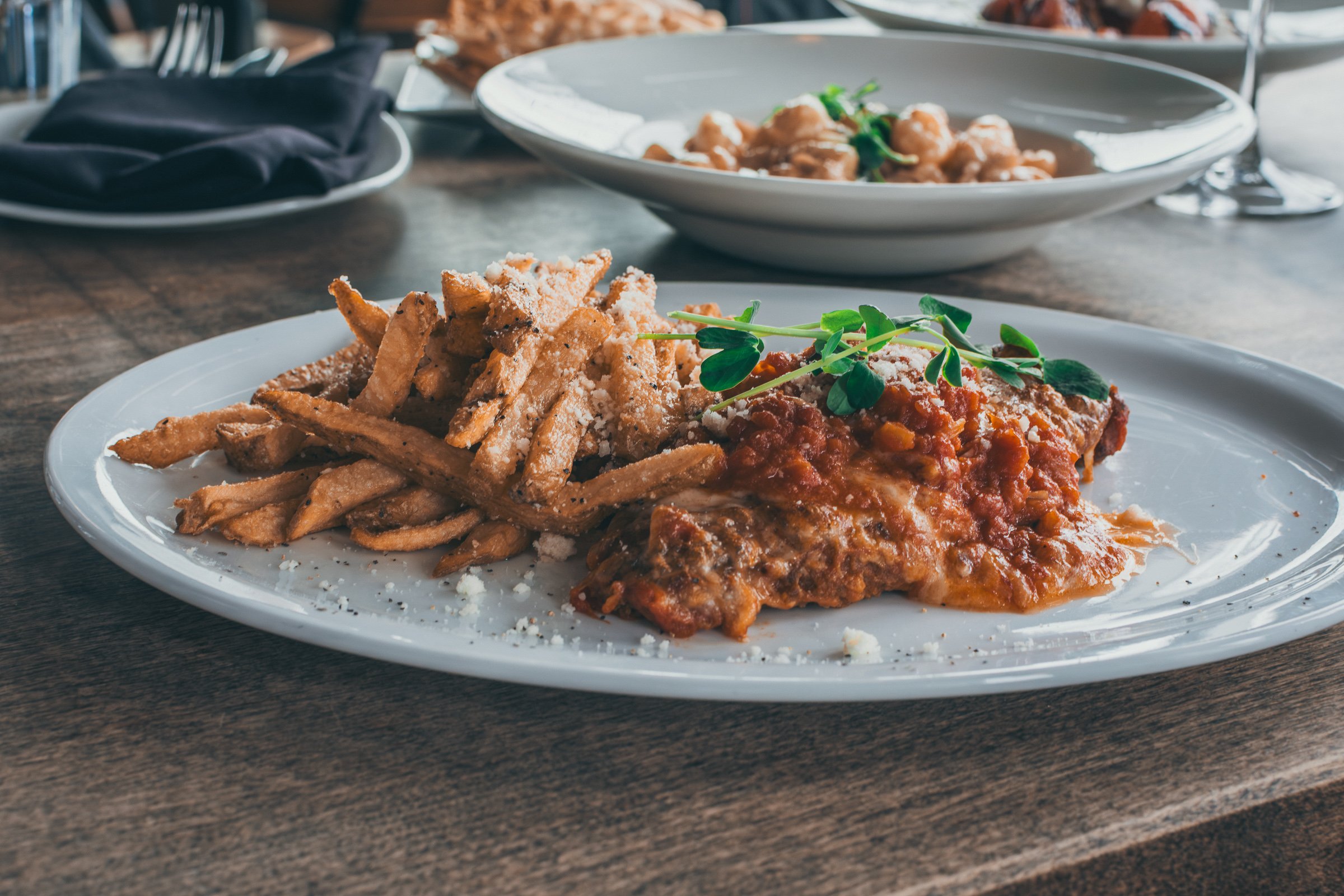 Plate of chicken Parmesan with spaghetti and garnished with green herbs, served on a white dish on a wooden table. In the background, to-go plates of food and a black napkin are visible.