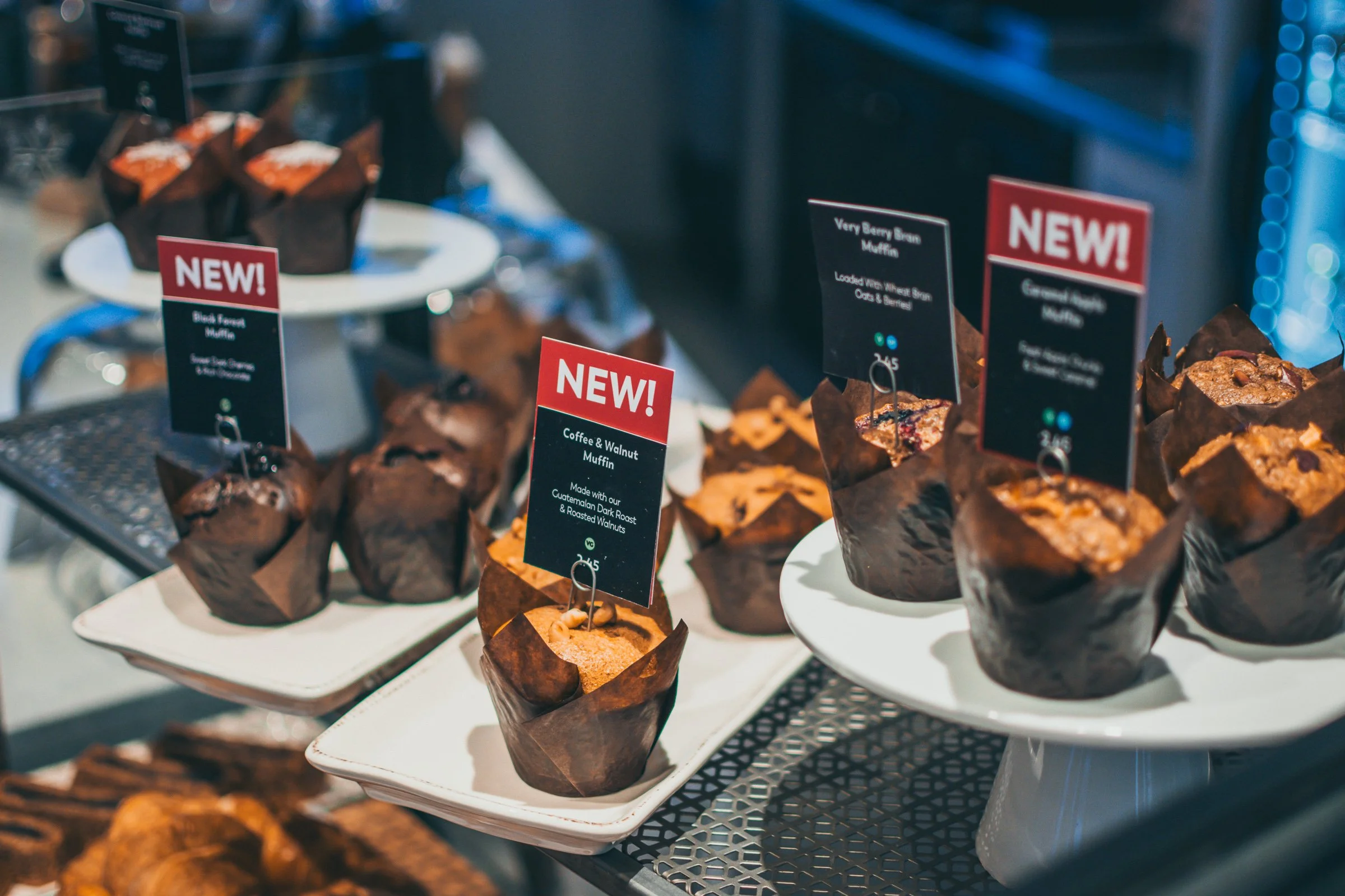 Display of muffins on a tiered tray with small signs labeled 'NEW.' The muffins are wrapped in brown paper, and the signs indicate flavors such as 'Black Forest Muffin,' 'Coffee & Walnut Muffin,' and 'Very Berry Bran Muffin.'