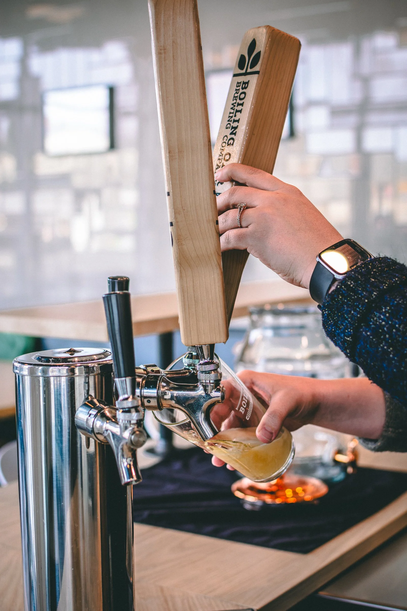 Person filling a glass with beer from a tap at a bar or brewery, with a wooden board labeled 'Brewing' being held over the glass.