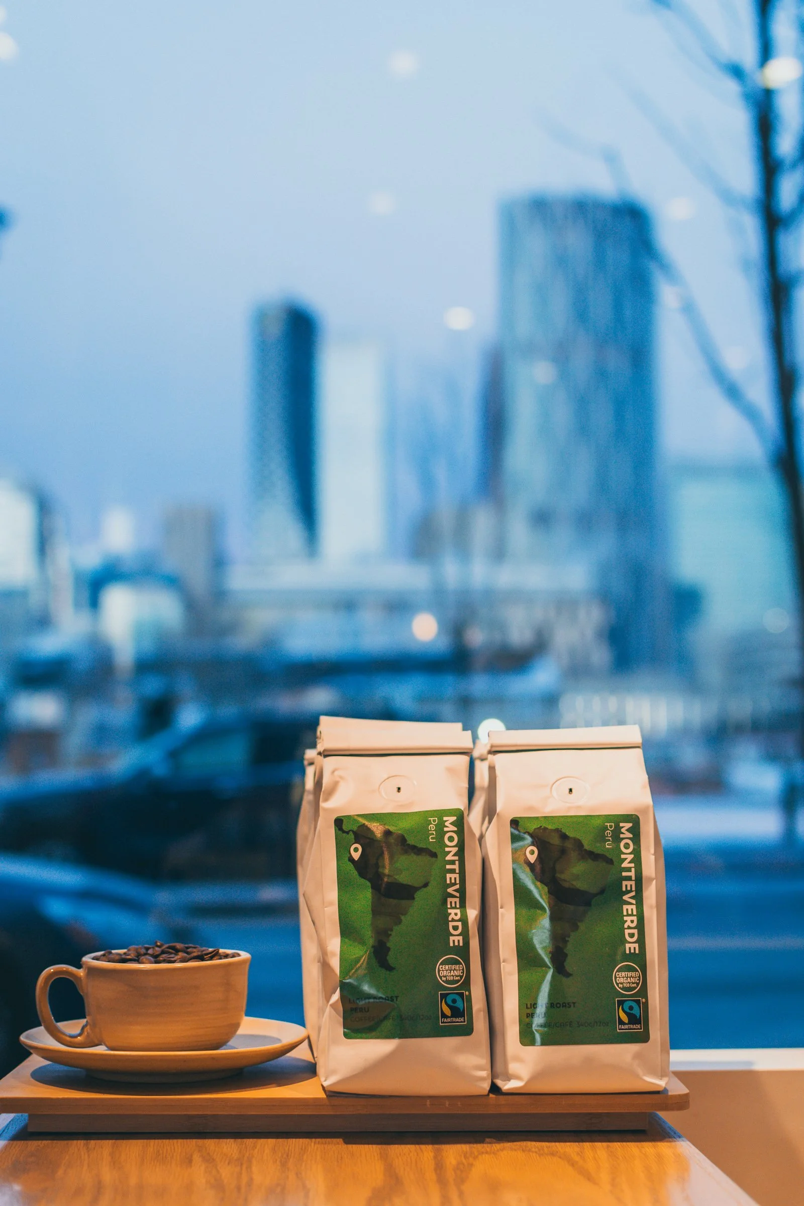 Two white bags of Monteverde coffee on a wooden table, with a cup of coffee beans on a saucer next to them, and a large window with city skyscrapers in the background.