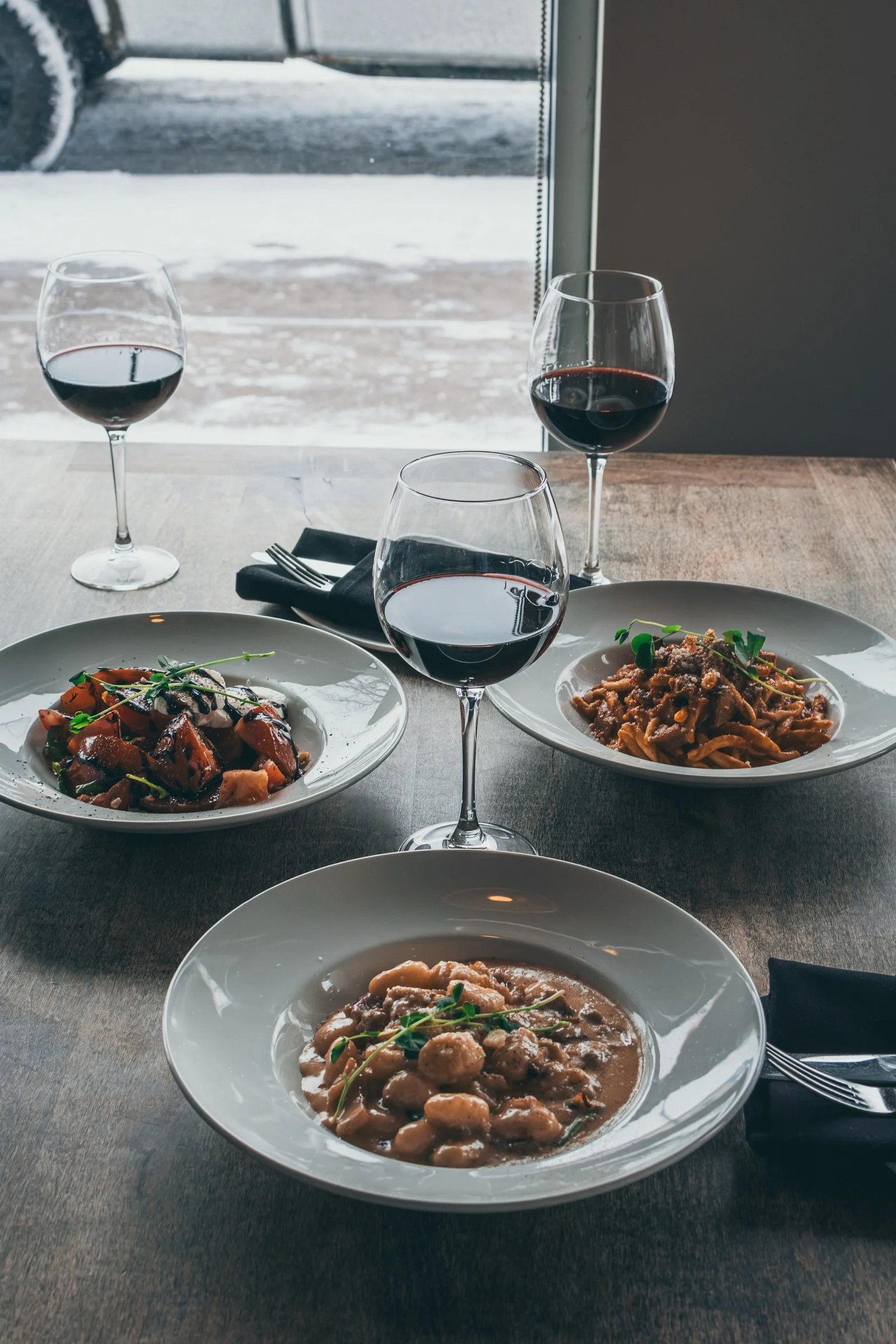 A table set with three plates of different cooked dishes, three glasses of red wine, and a black cloth napkin with a fork. Outside the window, snow covers the ground and a vehicle is visible.