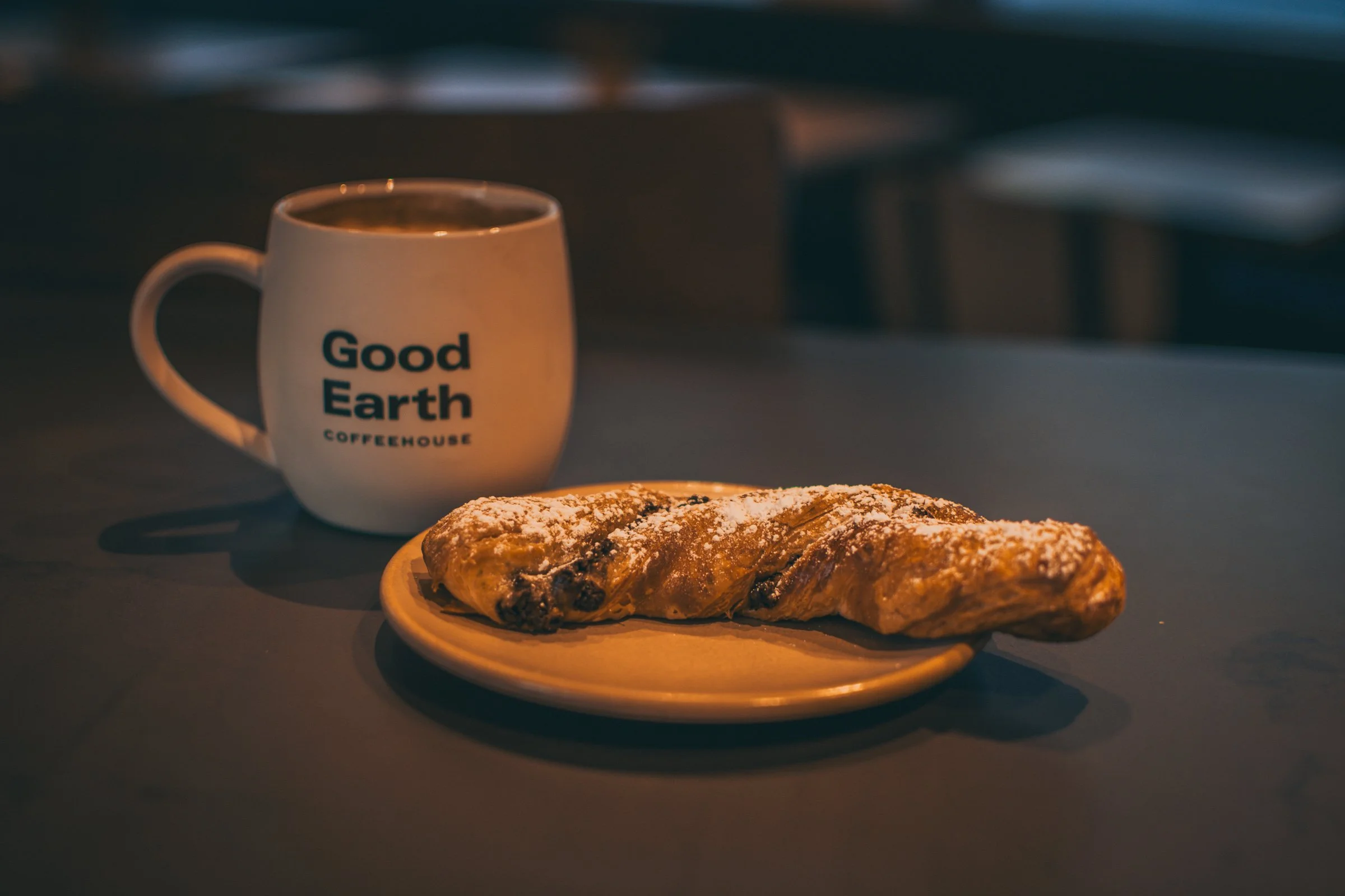 A white mug with the words 'Good Earth Coffeehouse' on it, placed next to a plate with a pastry dusted with powdered sugar on a dark surface. The background is blurred.