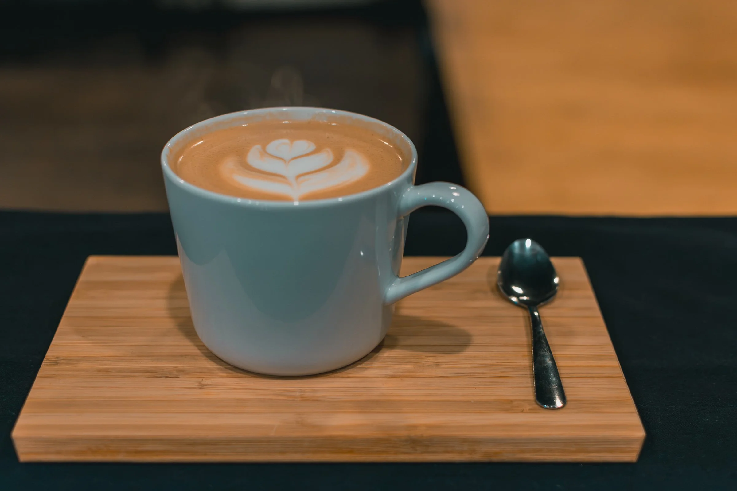 A white ceramic mug filled with a latte featuring latte art in the shape of a heart. The mug is on a wooden tray with a silver spoon beside it. The background is blurred.