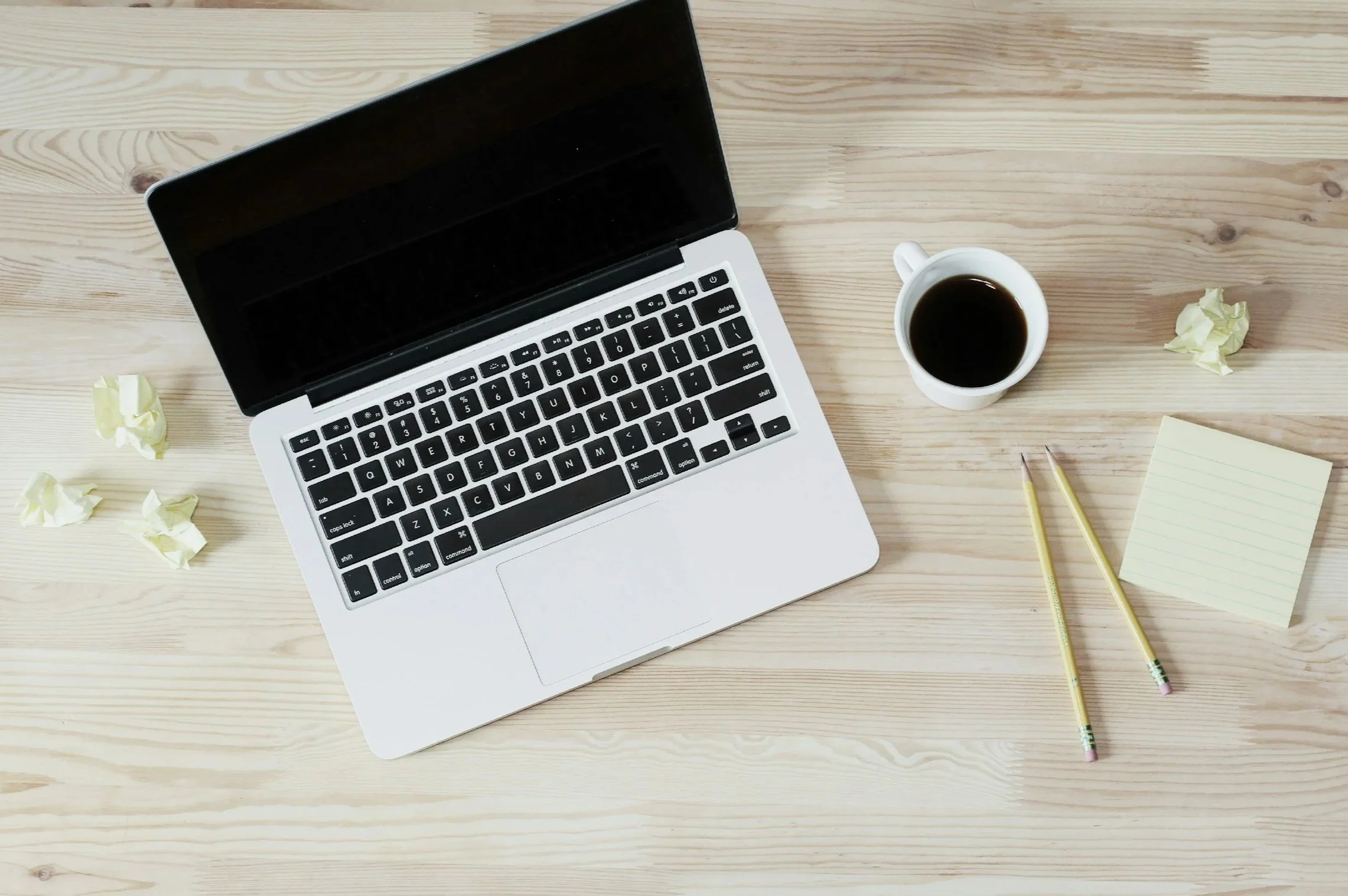 A laptop, a cup of coffee, two yellow pencils, a notepad, and crumpled pieces of paper on a wooden desk.