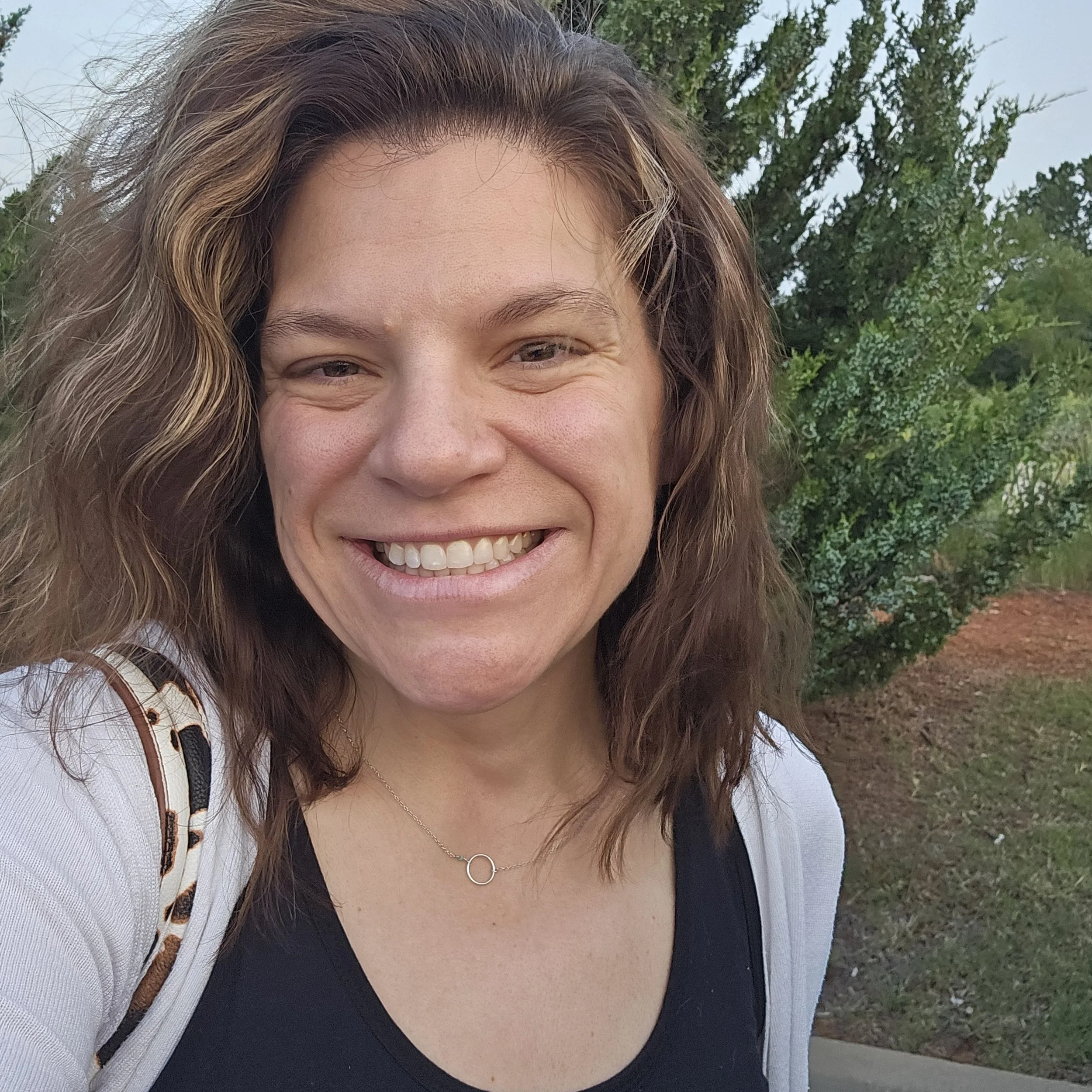 A smiling woman with brown, wavy hair outdoors, wearing a black top and a white cardigan, with trees and cloudy sky in the background.
