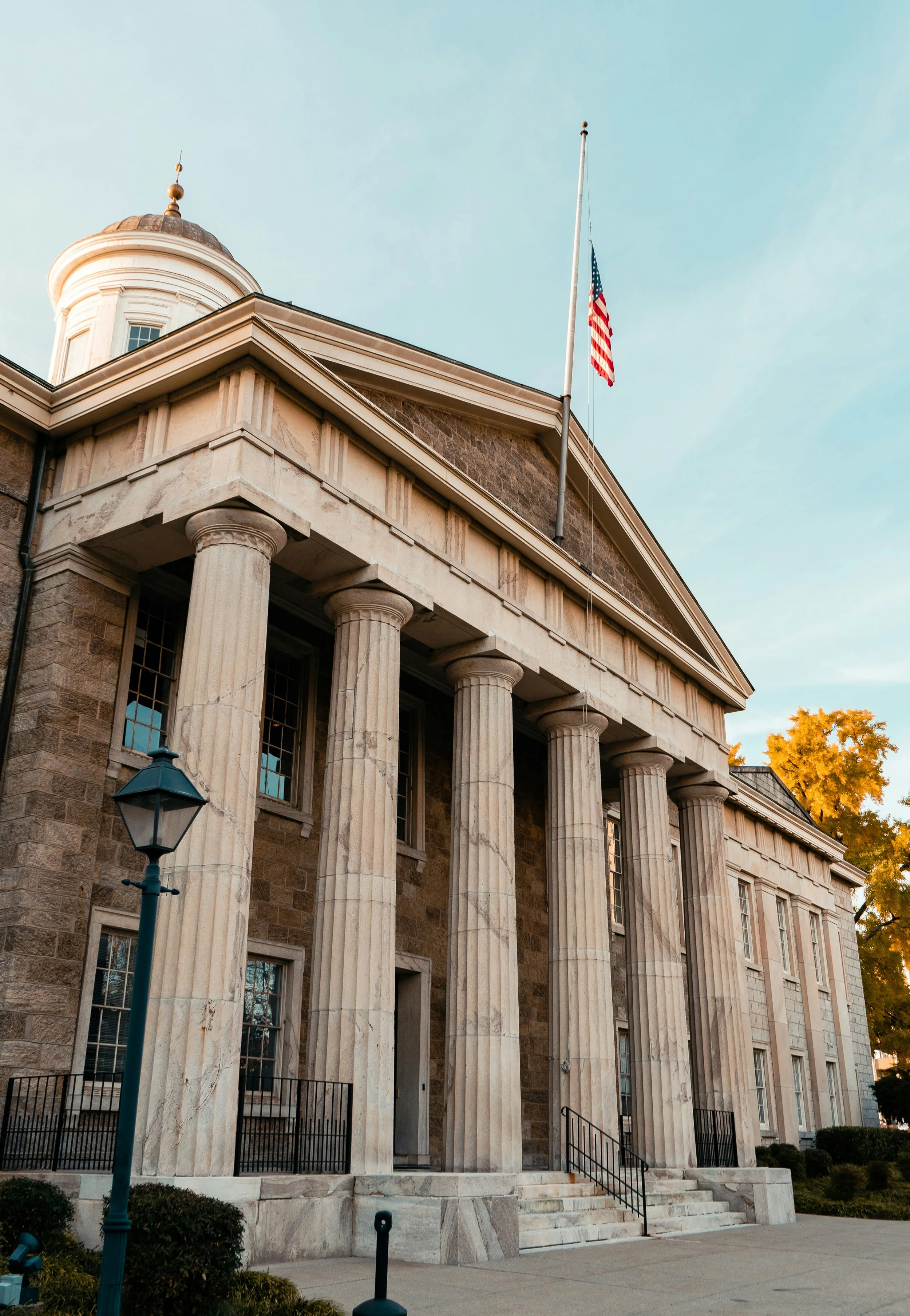 A historic courthouse building with large columns, stone steps, and an American flag flying on a flagpole in front.
