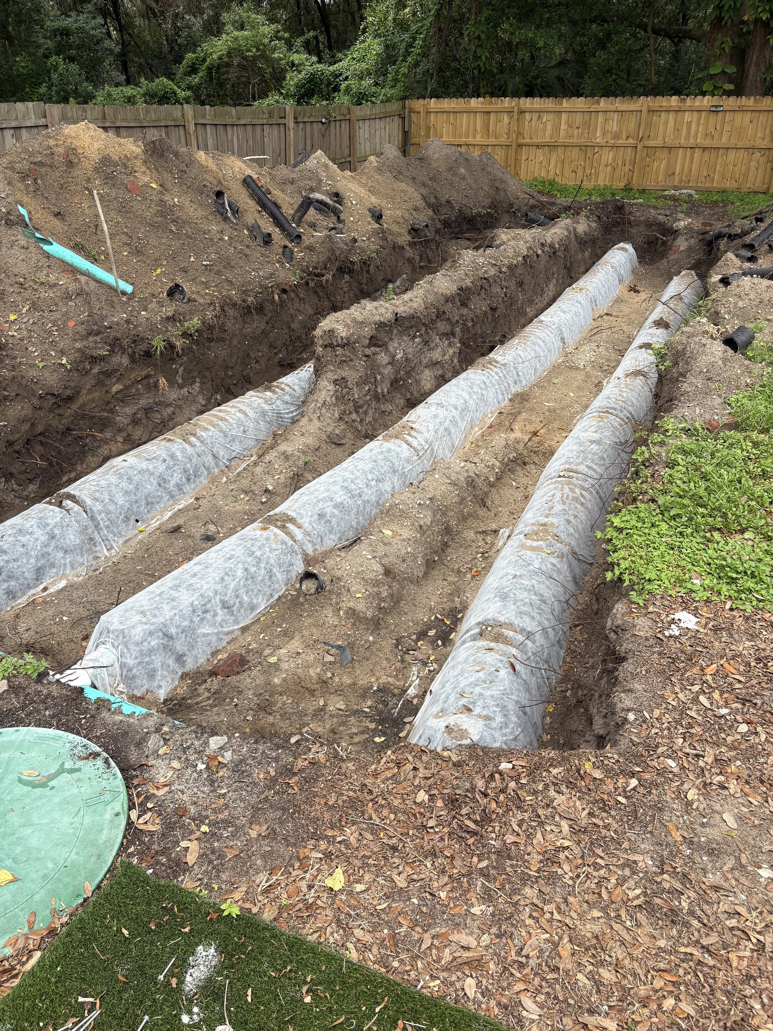 Underground plumbing installation with large black pipes and large insulated pipes in a backyard with a wooden fence, trees, and fallen leaves.