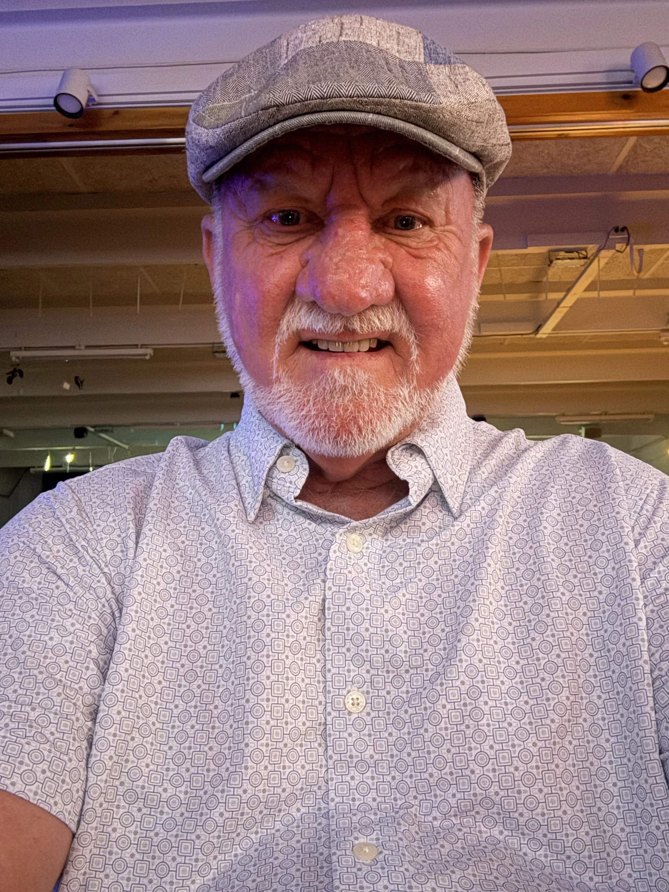 A close-up selfie of an elderly man with a white beard, wearing a patterned button-up shirt and a gray flat cap, in an indoor setting with ceiling lights and wood paneling.