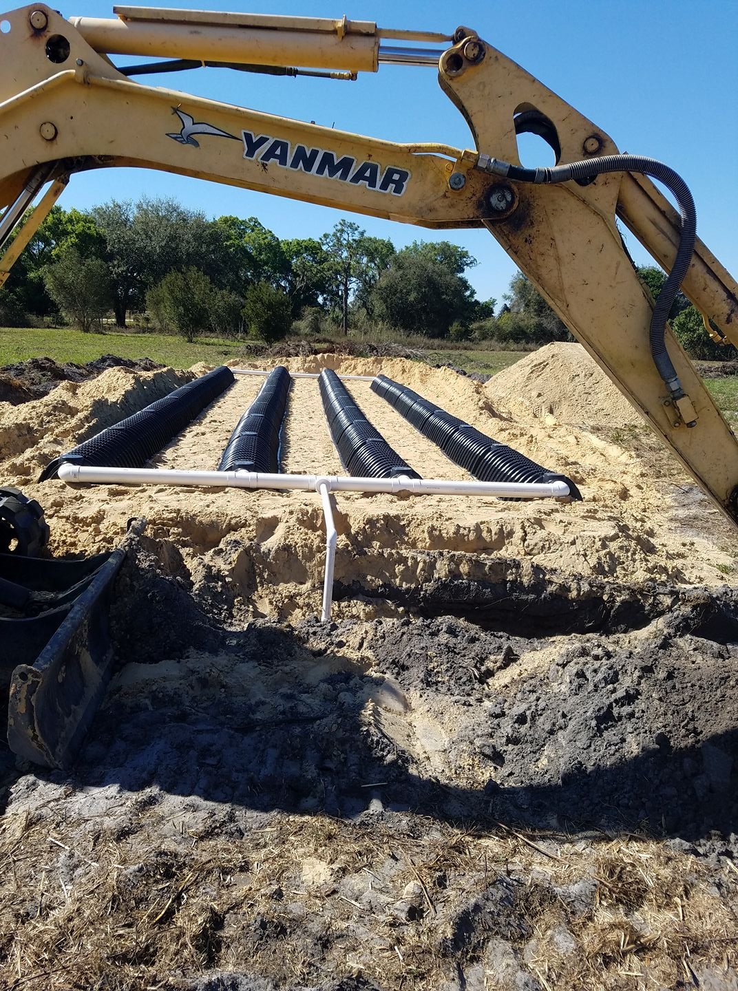 Drain pipes installation with pipes laid in a trench and a construction excavator in a field with trees and blue sky in the background.