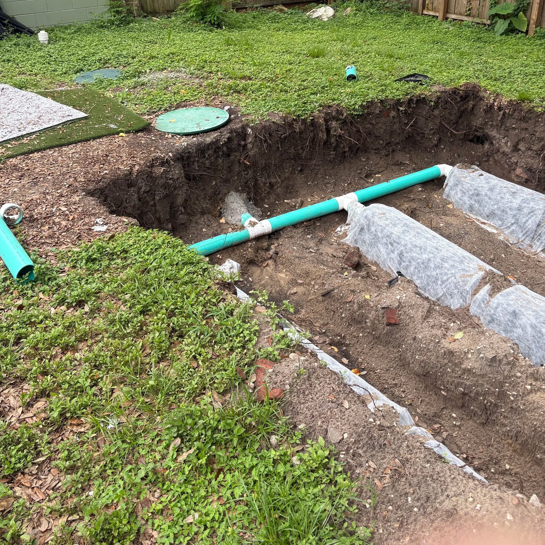 A backyard yard with grass and weeds, a wooden fence, a green pipe, and an open hole in the ground with weeds growing inside.