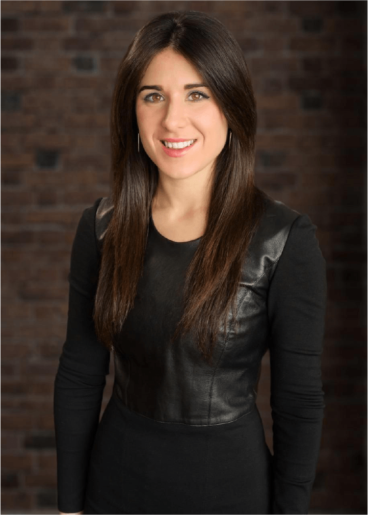 A woman with long brown hair, wearing a black top with leather panels, smiling, standing against a brick wall background.